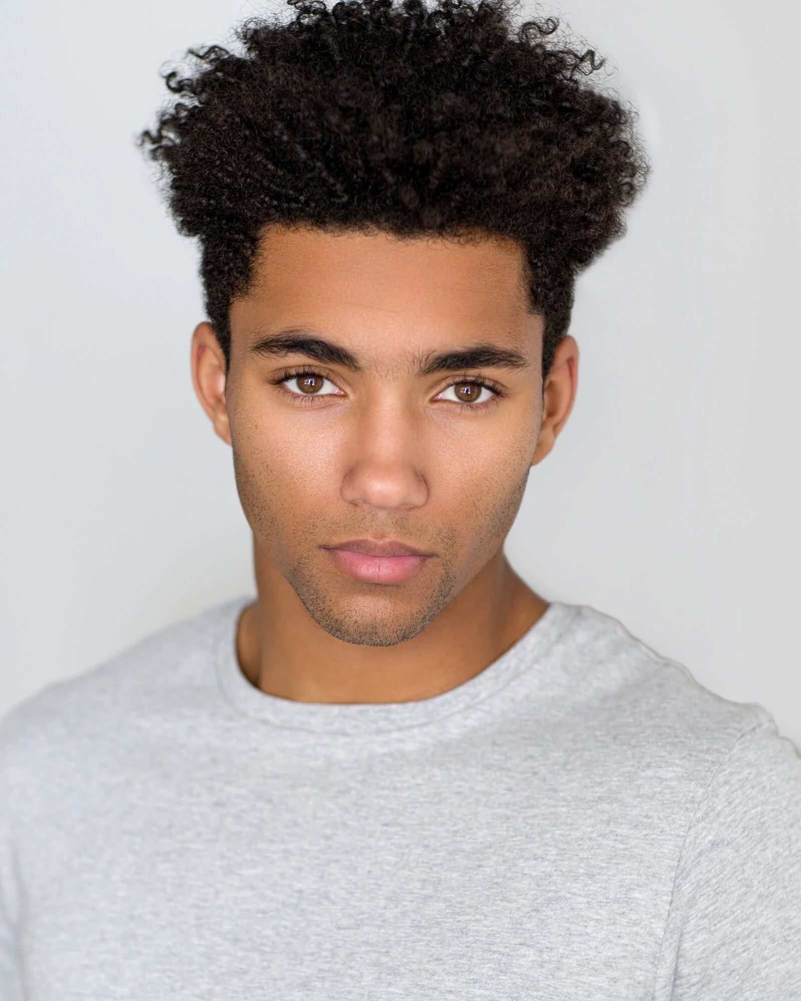 Headshot of a young man with curly dark hair and brown eyes, wearing a light gray shirt, looking directly at the camera against a plain white background.