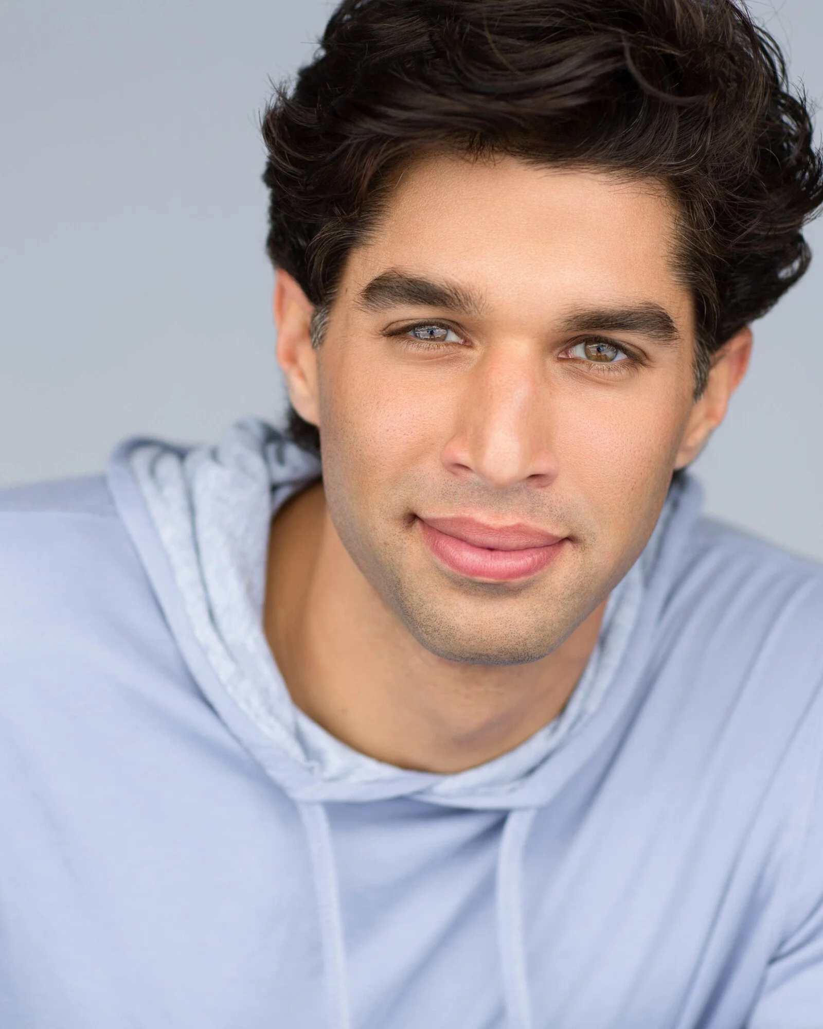 Close-up of a young man with dark, wavy hair, light blue eyes, and a light complexion, wearing a light gray hoodie with a drawstring.