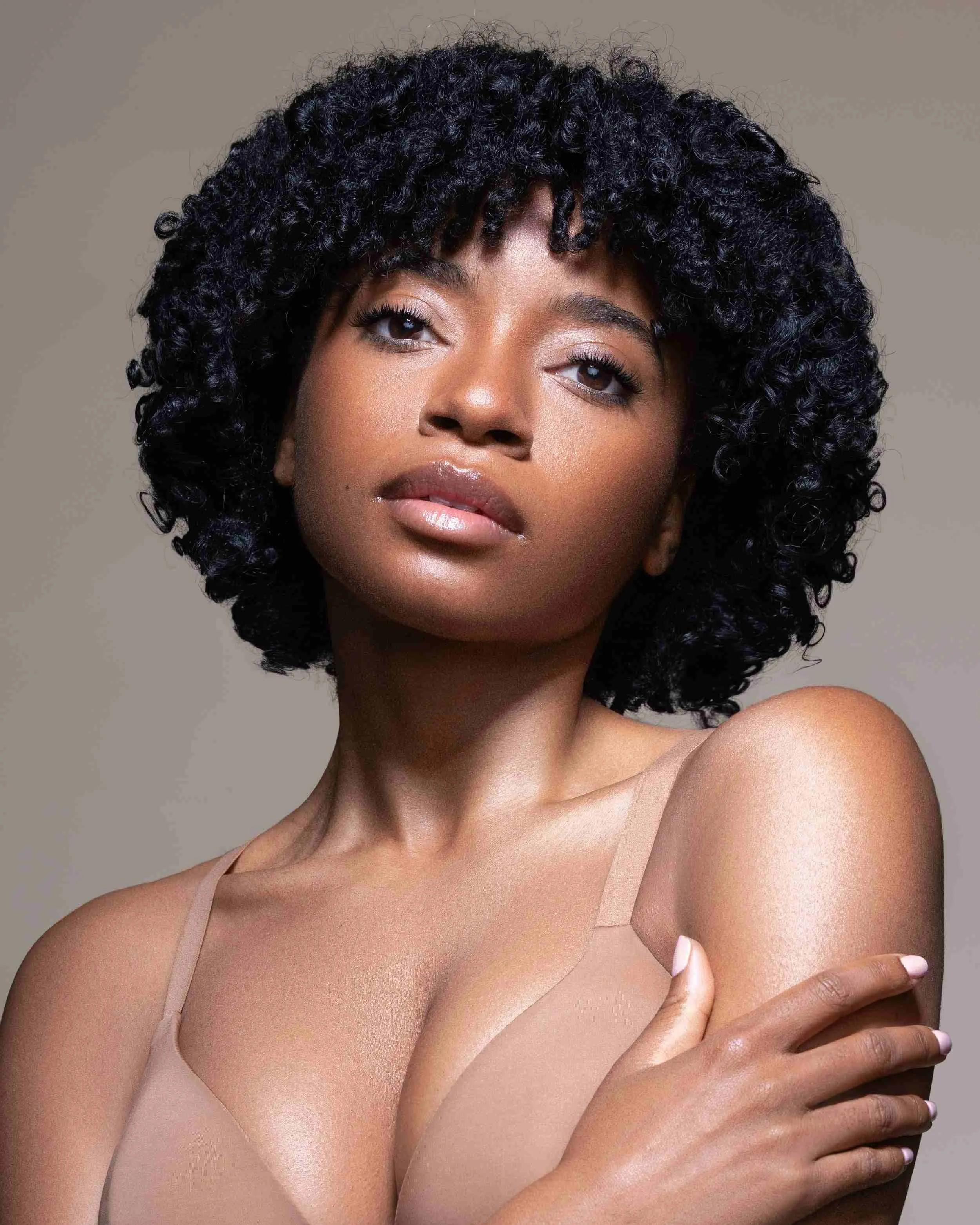 A young woman with curly black hair and brown skin poses with her hand on her shoulder, wearing a beige bra, against a neutral background.