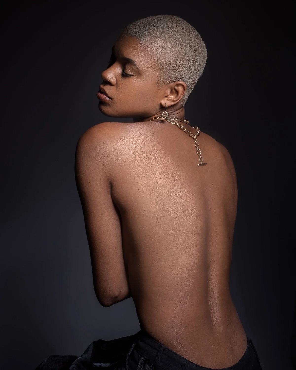 A woman with short blonde hair and dark skin poses with her back to the camera and eyes closed against a dark background. She is topless, wearing a chunky gold chain necklace and earrings.