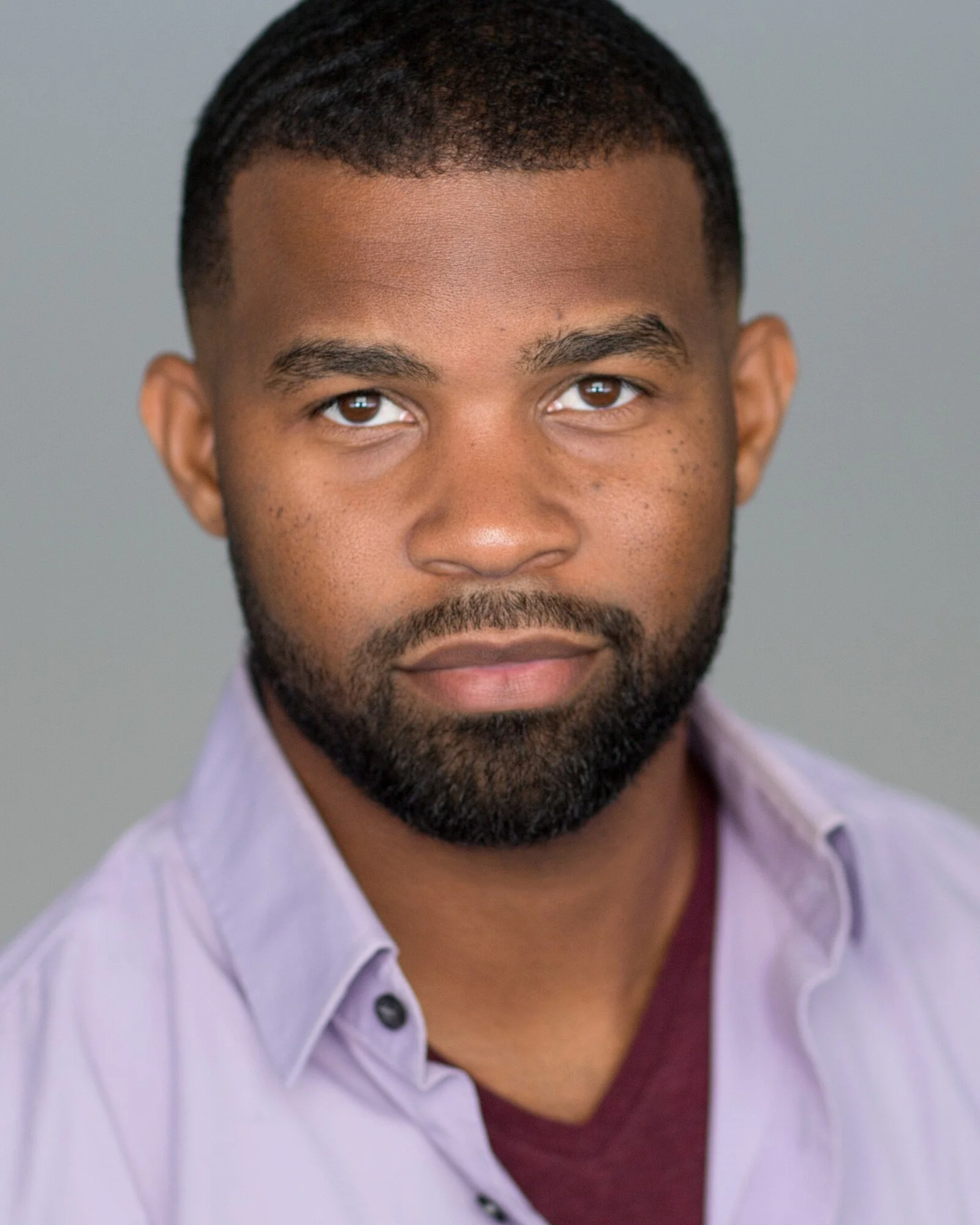 Close-up of a serious African American man with a beard and short hair, wearing a light purple button-up shirt with a maroon shirt underneath, against a plain gray background.