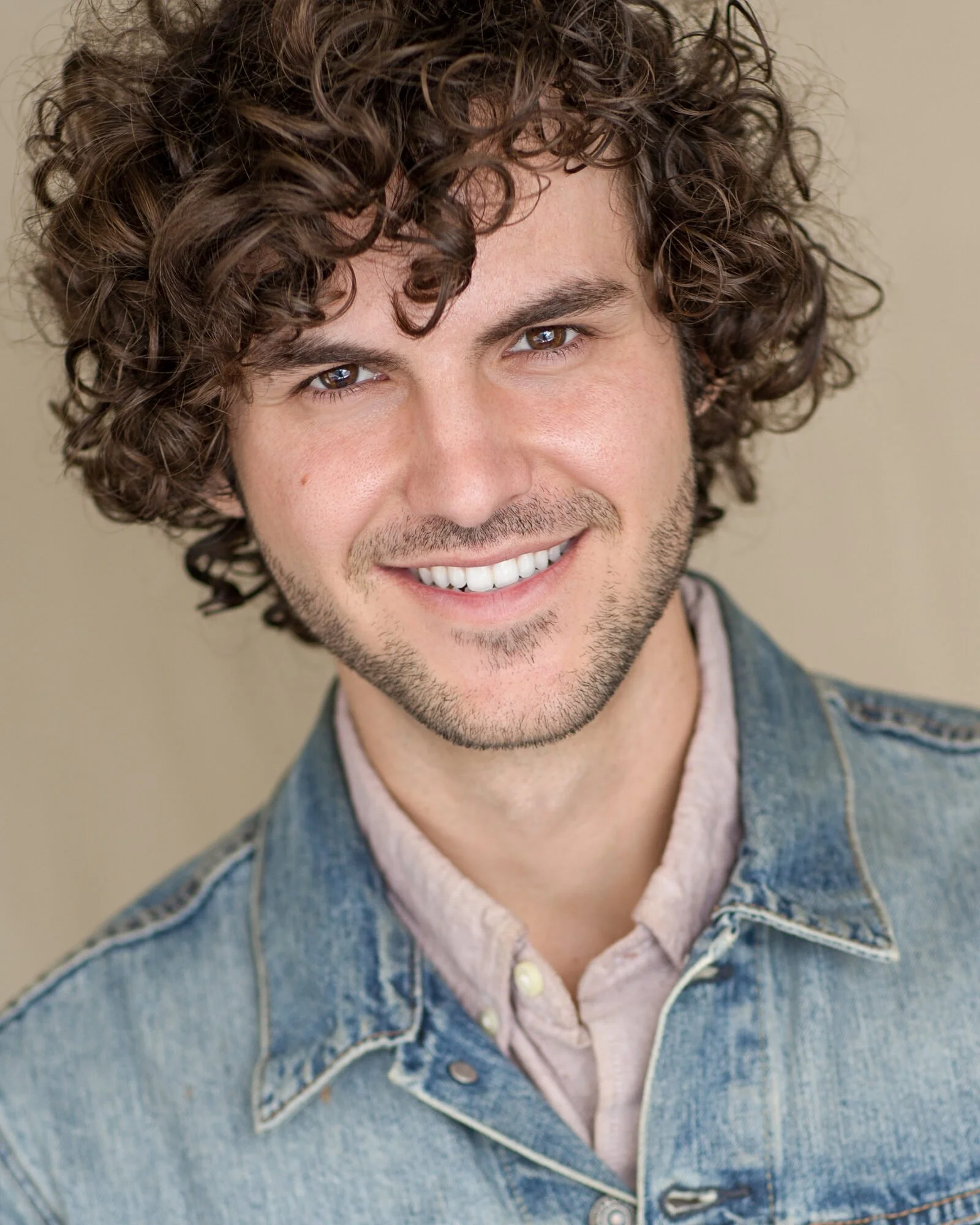 A young man with curly brown hair, brown eyes, and a light beard, smiling, wearing a denim jacket over a light pink shirt, against a neutral background.