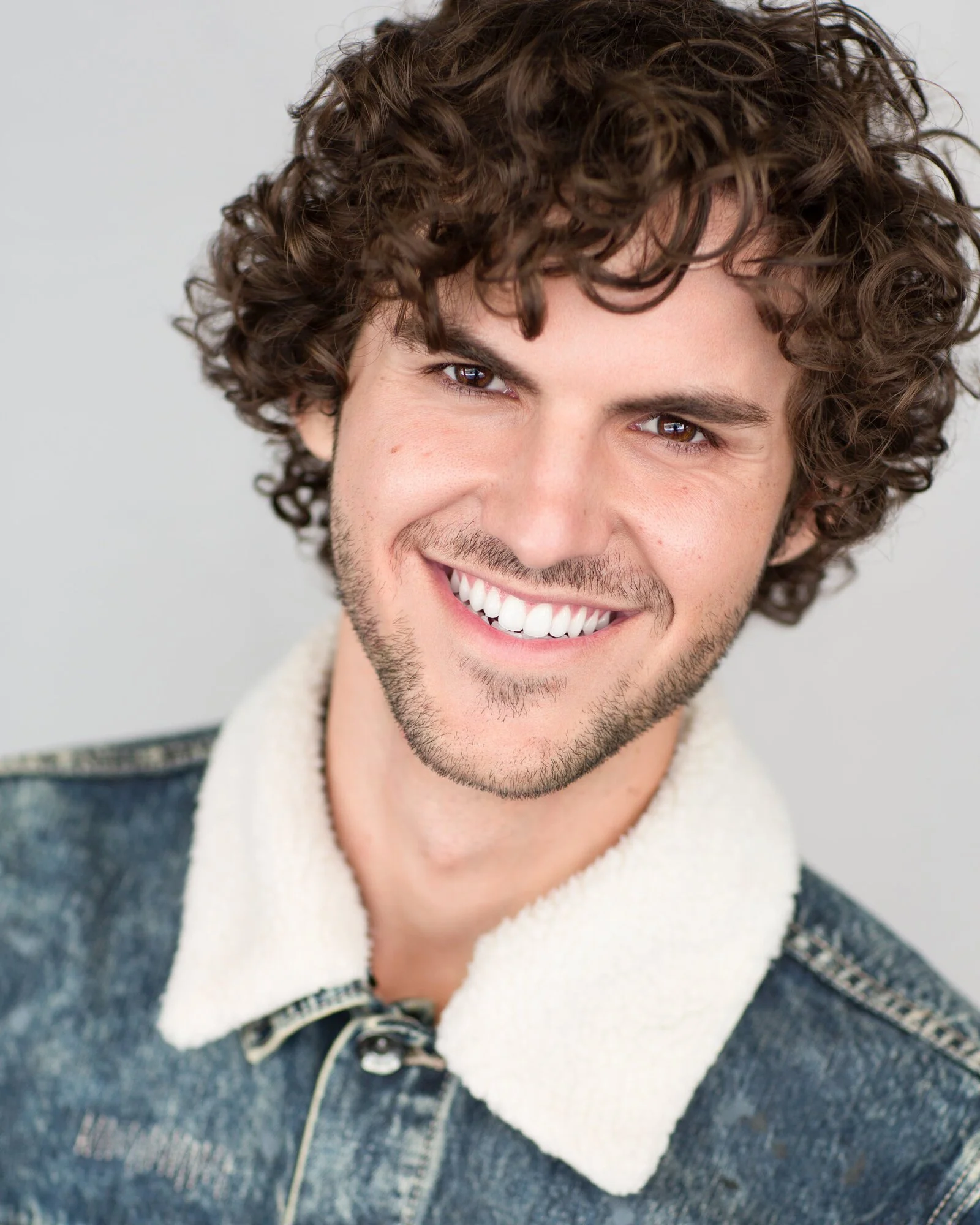 A young man with curly brown hair, brown eyes, and a bright smile, wearing a denim jacket with a white fleece collar.