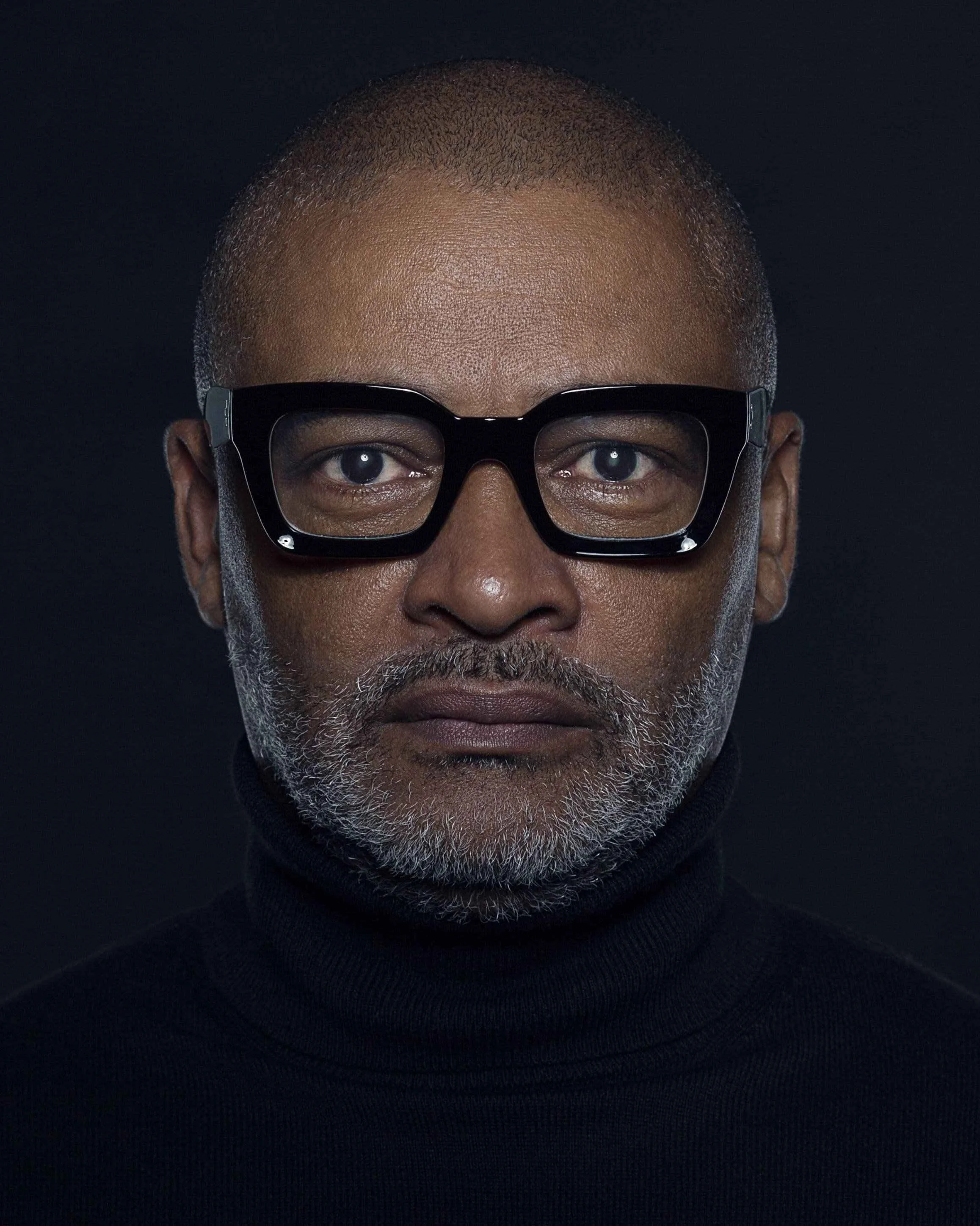 A mature Black man with glasses, gray beard, wearing a black turtleneck, looking directly into the camera against a dark background.
