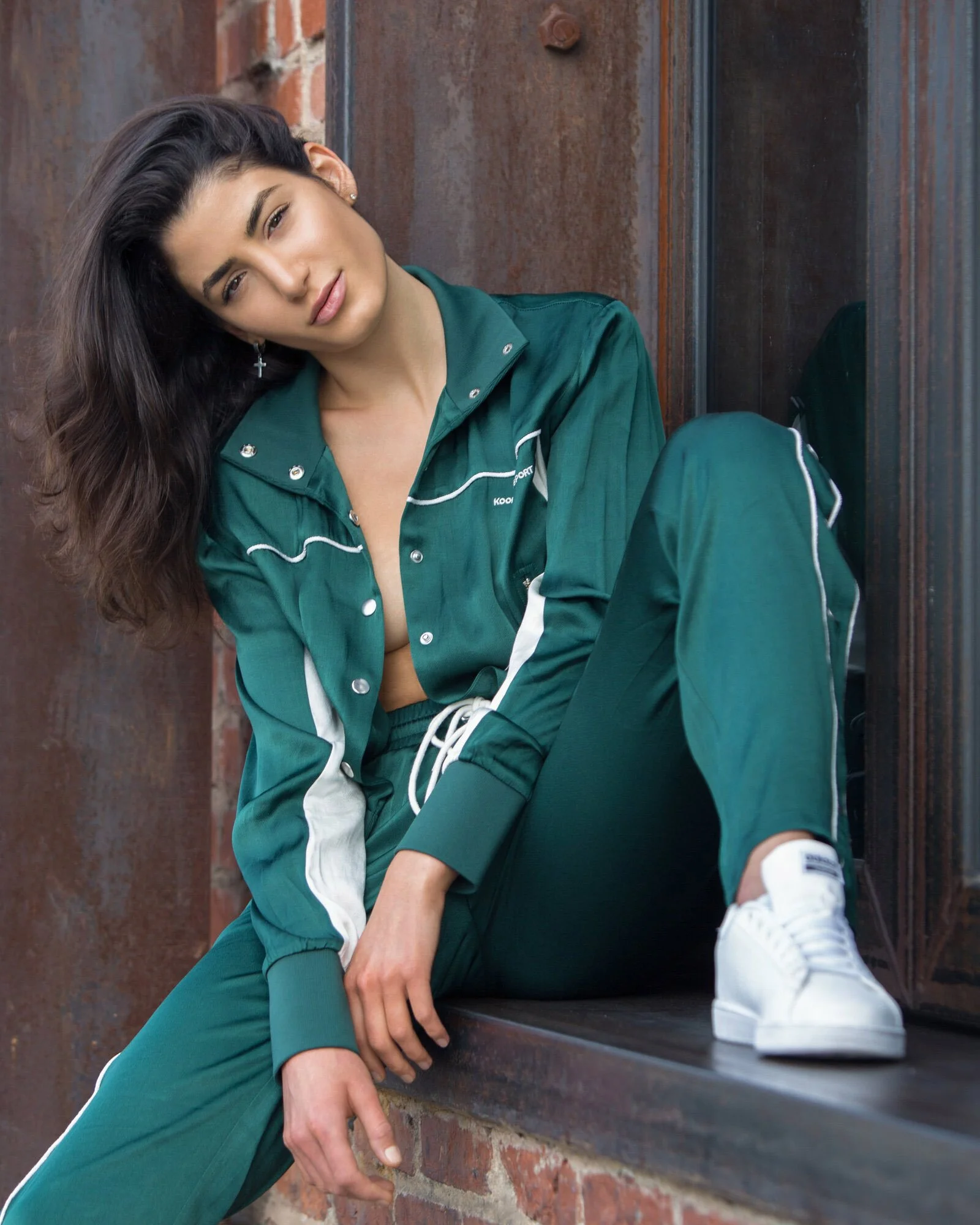 A young woman with long dark hair, wearing a green athletic tracksuit and white sneakers, sitting on a brick windowsill next to a rusty metal wall.