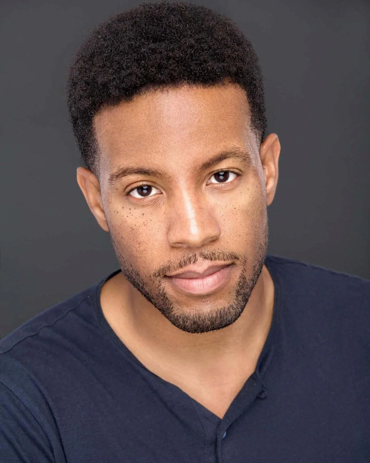 Close-up portrait of a young Black man with short curly hair and a slight beard, wearing a dark blue shirt, against a plain dark background.