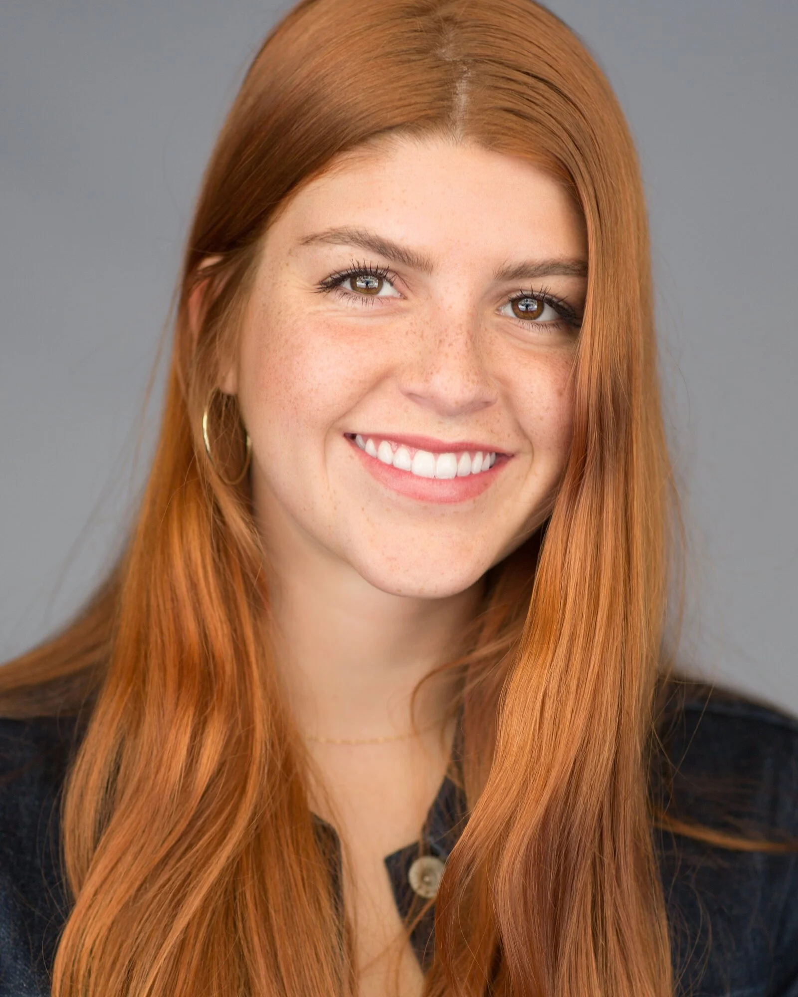 A young woman with long red hair, brown eyes, and freckles, smiling at the camera, wearing a black top and hoop earrings against a gray background.