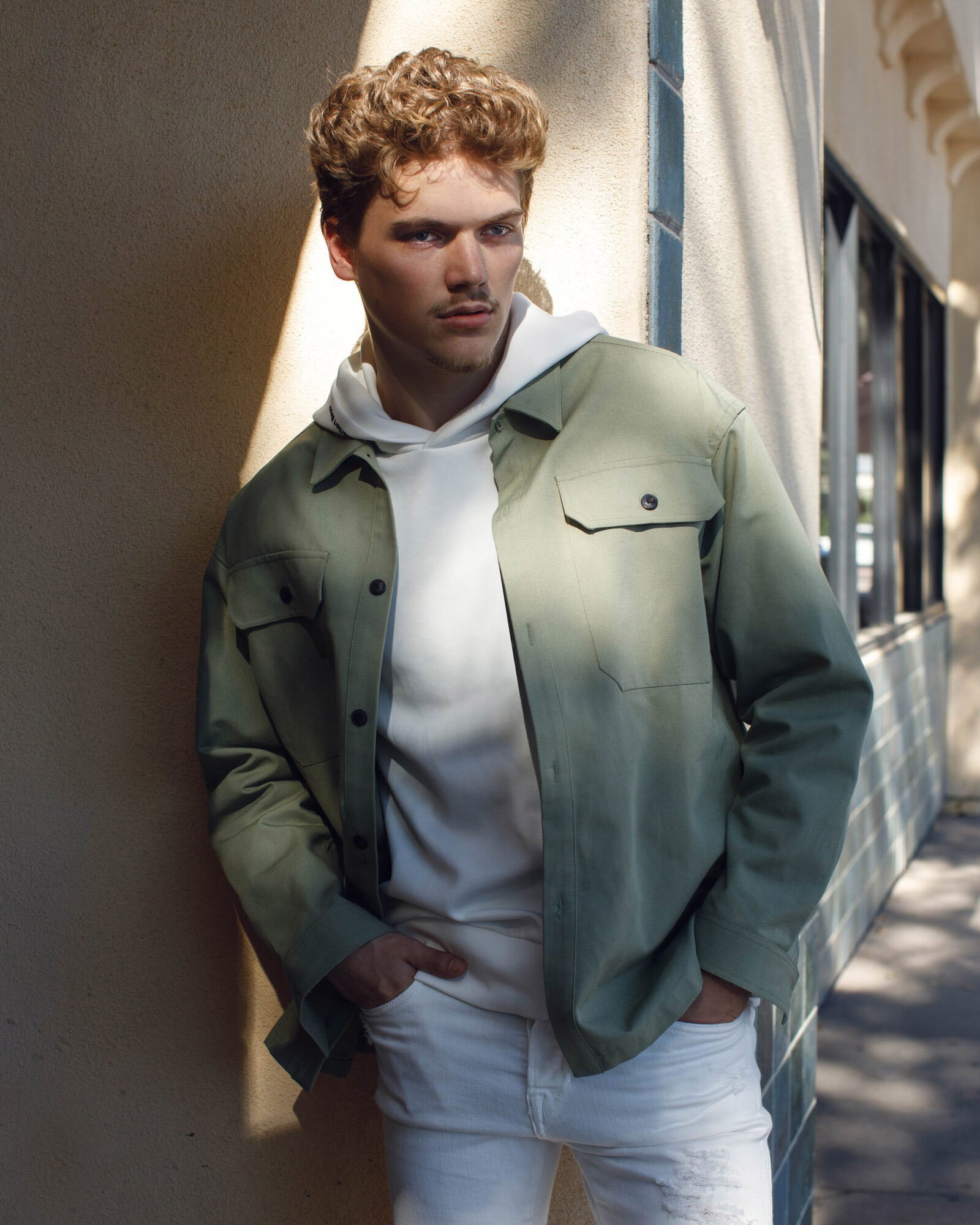 A young man with curly hair leaning against an outdoor wall, wearing a light green jacket over a white hoodie and white pants, with hands in pockets.