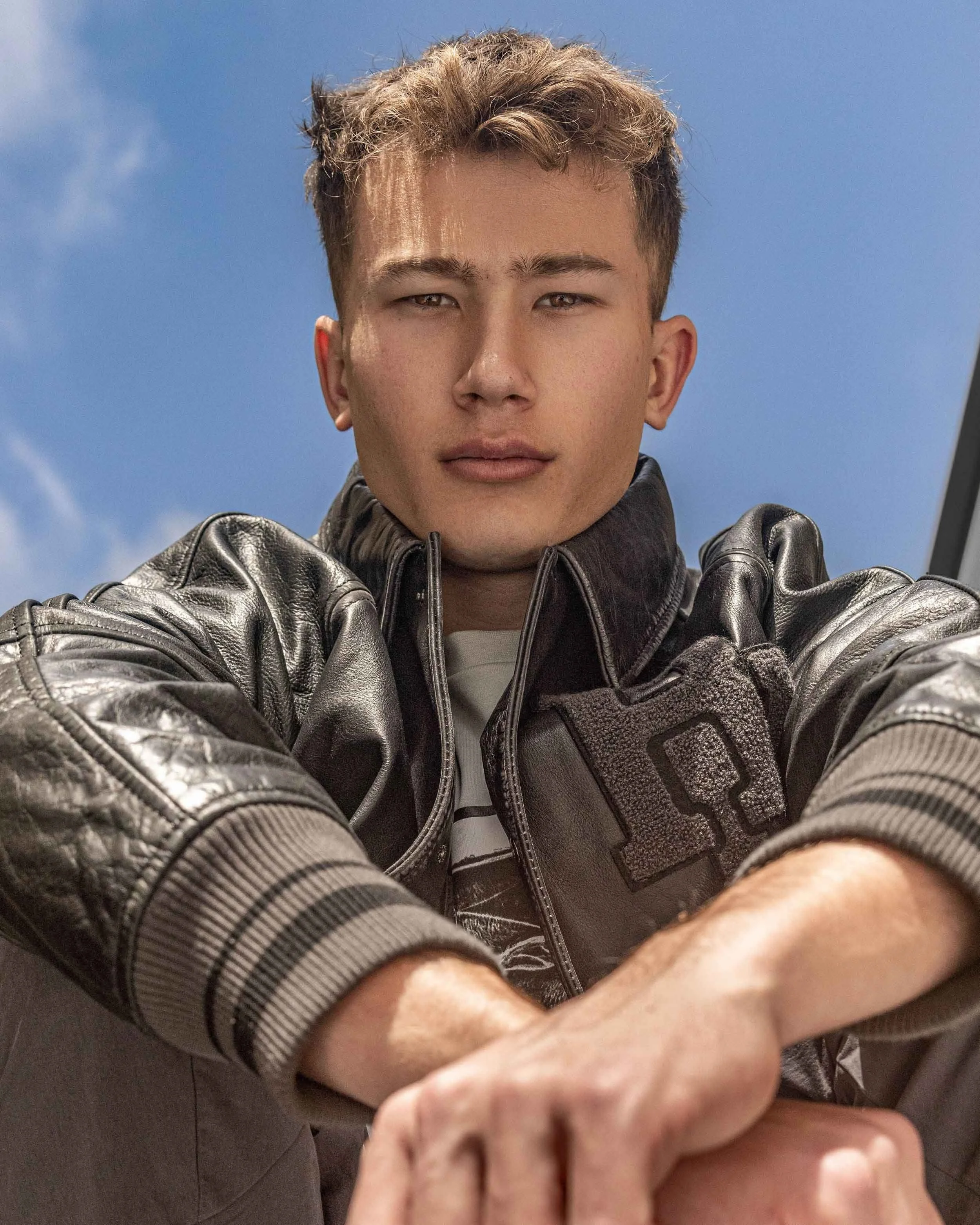 Young man with short brown hair wearing a black leather jacket and sitting outdoors against a blue sky.