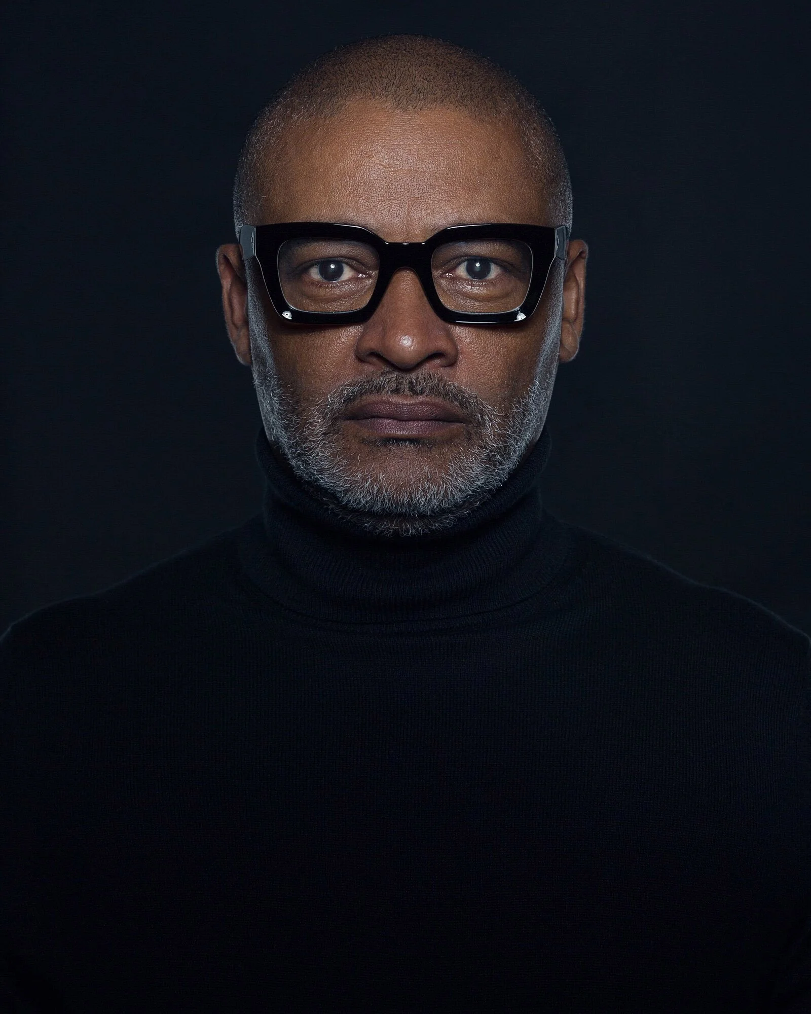 A close-up portrait of a middle-aged man with glasses, a gray beard, and a black turtleneck against a dark background.