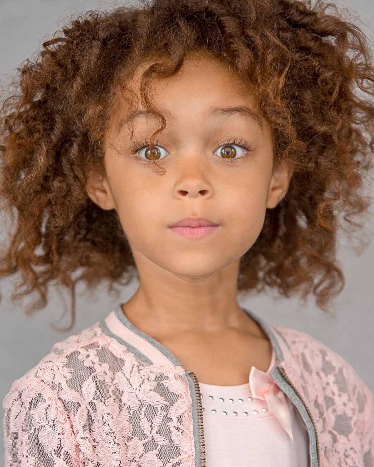 Close-up of a young girl with curly brown hair, brown eyes, and a neutral expression, wearing a pink lace jacket over a white top with a pink bow and small rhinestones.