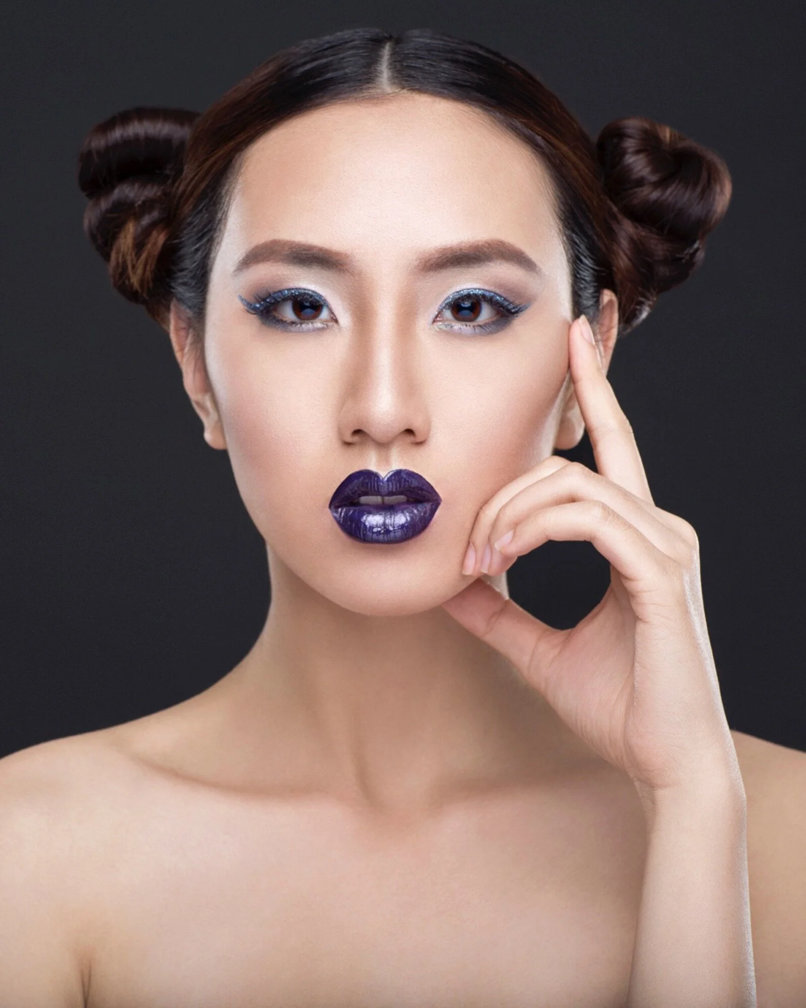 A woman with styled brown hair, wearing bold purple lipstick, glittery blue eye makeup, and posing with her hand near her face against a dark background.