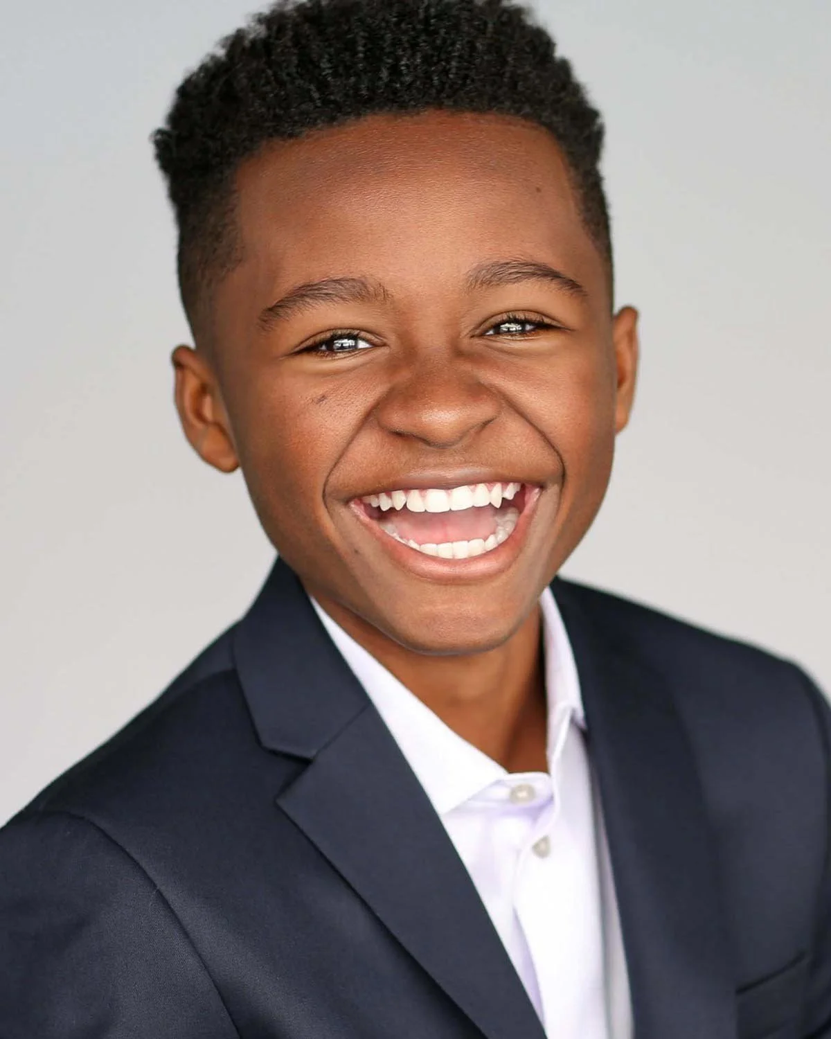 Smiling young boy in a dark suit and white shirt, facing forward against a plain background.