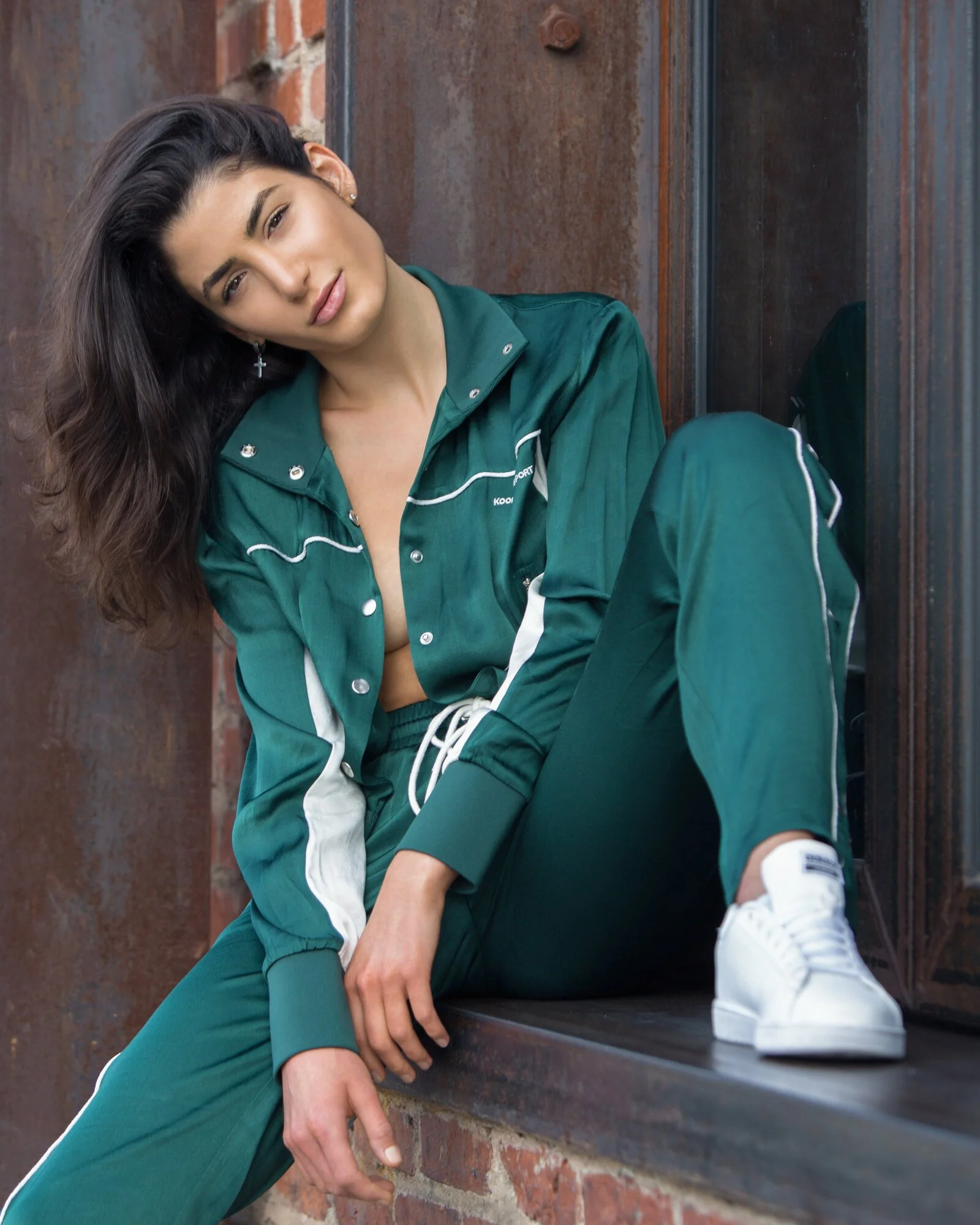 A young woman with dark wavy hair, wearing a green sports jacket and matching pants, sitting on a brick ledge with a rusted metal wall behind her.