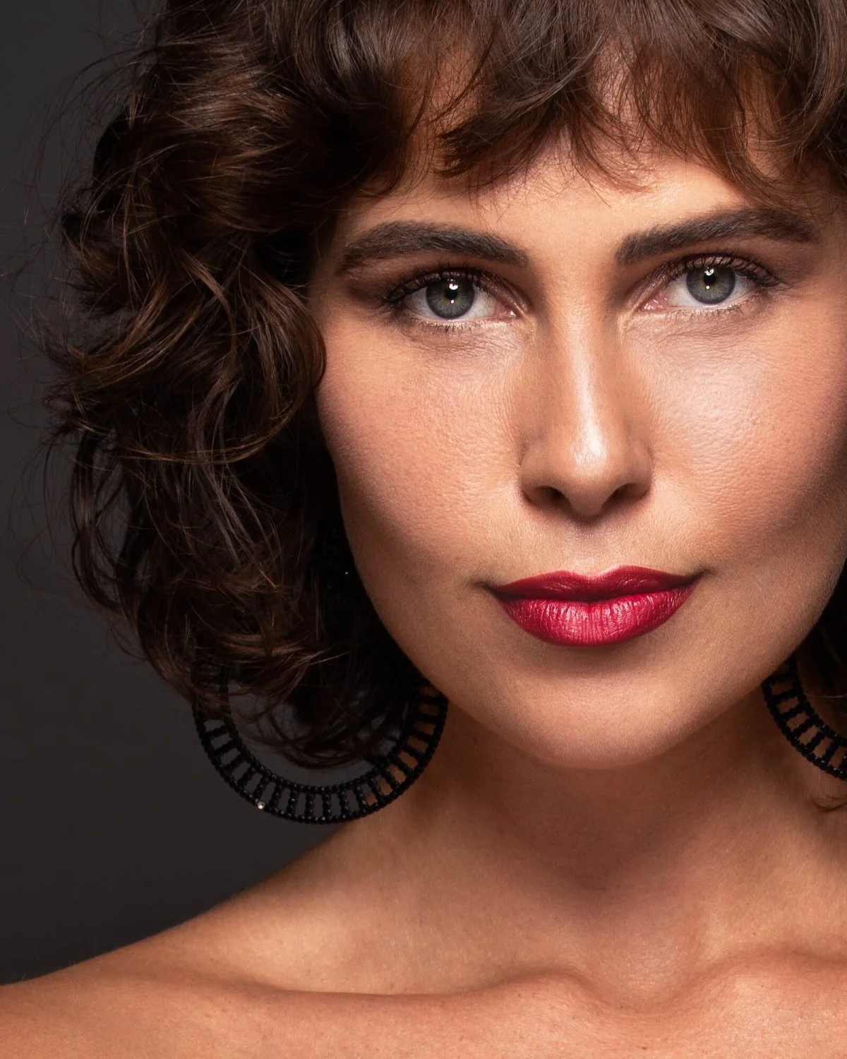 Close-up of a woman with short, curly brown hair, blue eyes, red lipstick, and black circular earrings, against a dark background.