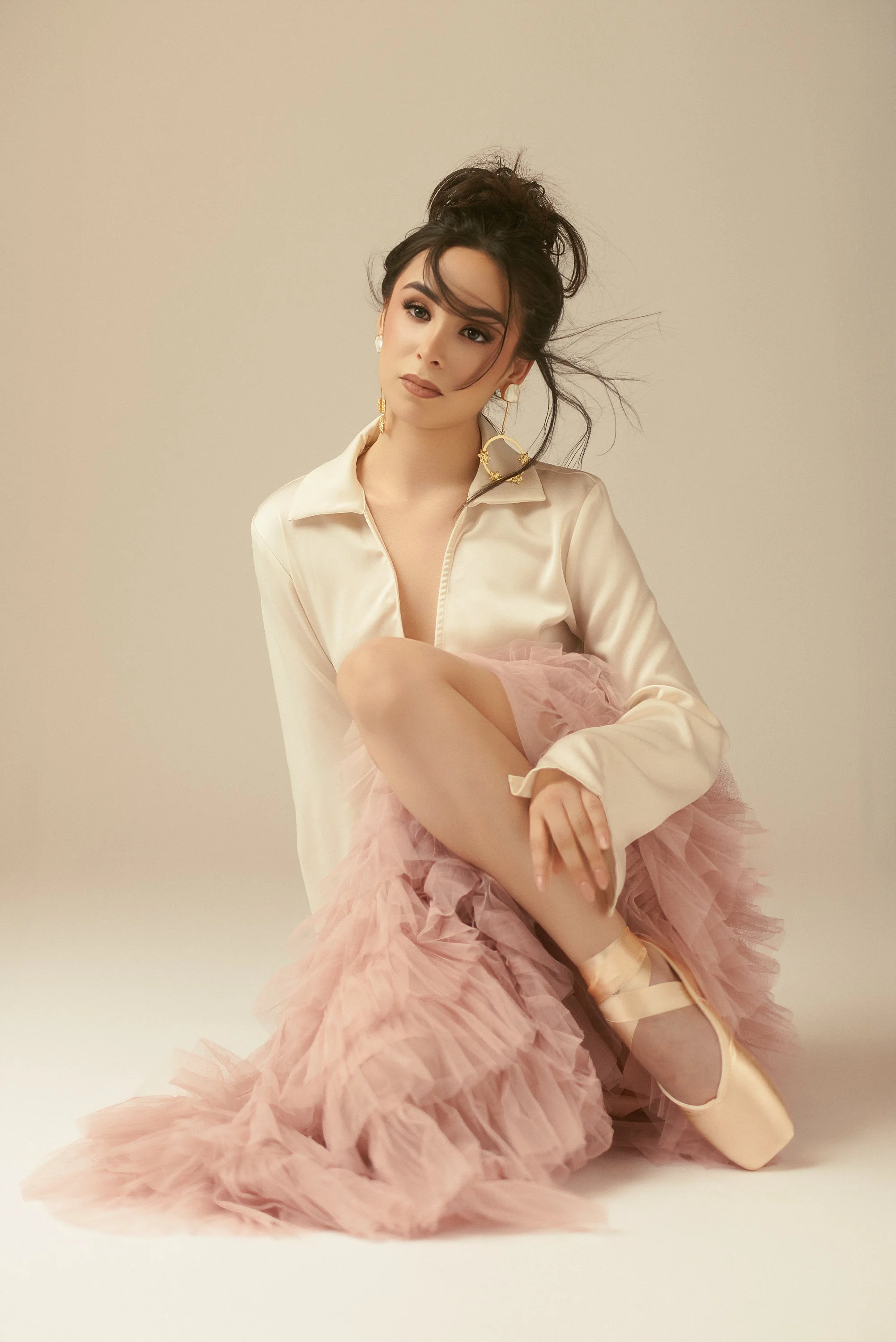 Una mujer posando con tutú rosa y zapatillas de ballet,piel clara, cabello oscuro y rizado, usando una blusa de satén clara y pendientes grandes de aro dorados.