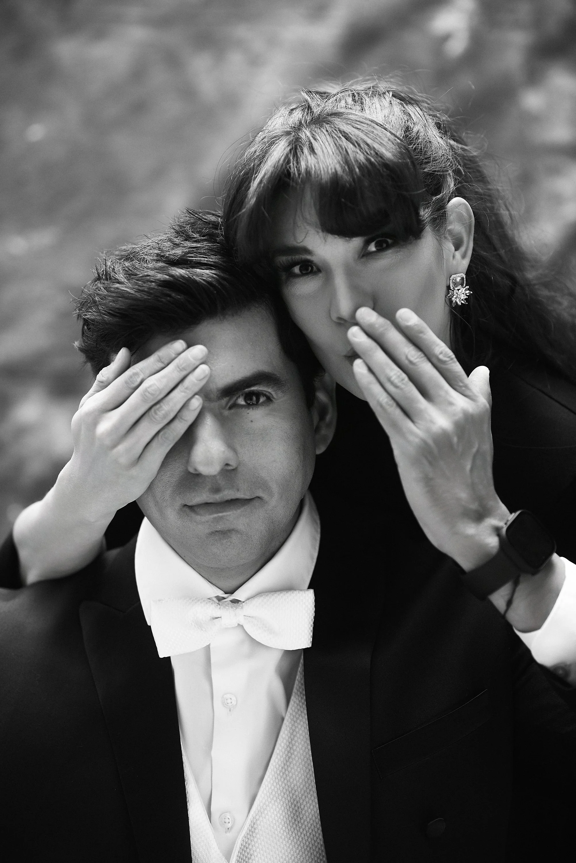 Retrato en blanco y negro de un hombre con ropa formal y una mujer que lo abraza por encima, ambos cubriéndose parcialmente el rostro con las manos, en un fondo difuso.