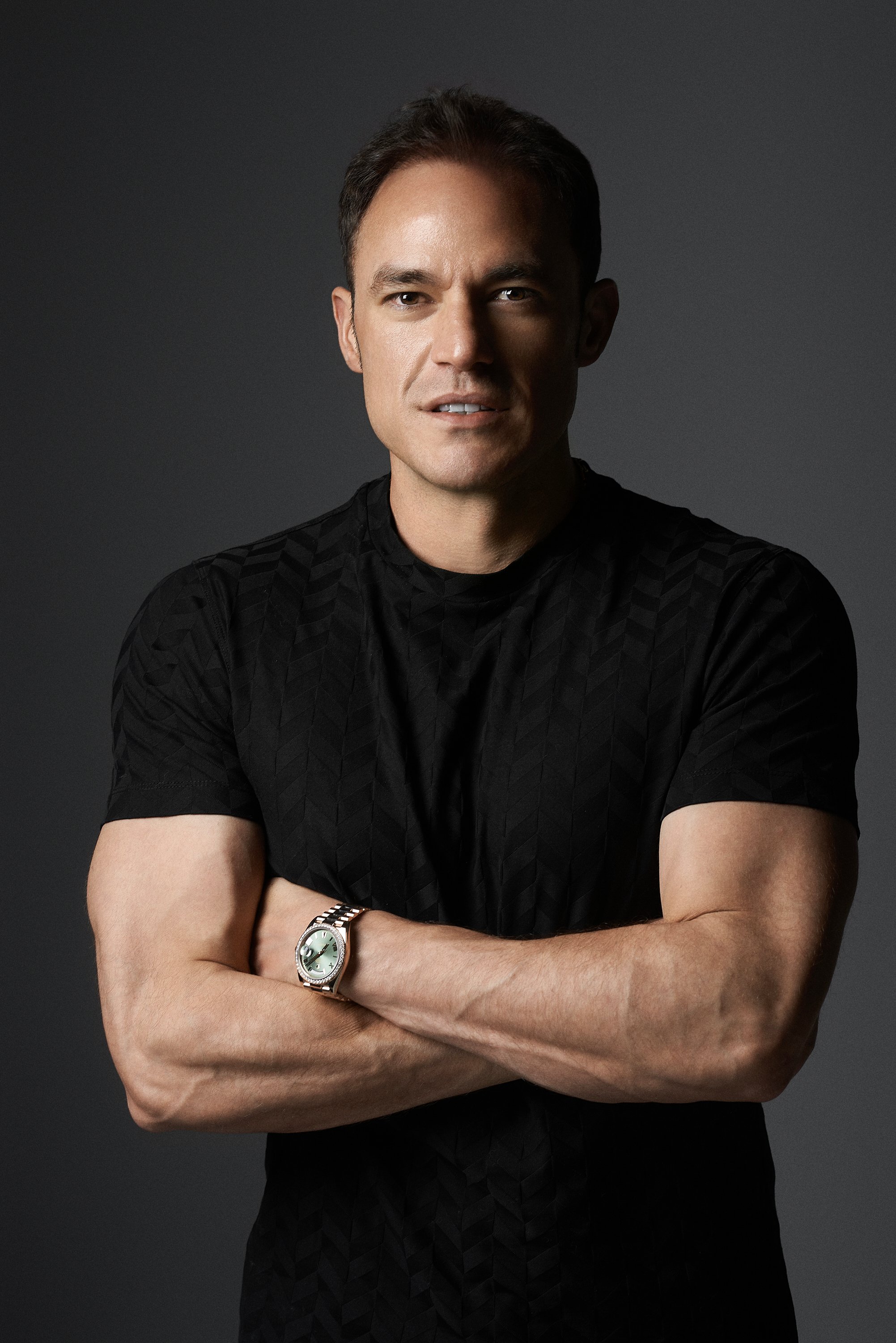 Retrato de un hombre con camiseta negra, brazos cruzados, luciendo un reloj en su muñeca izquierda, fondo oscuro.