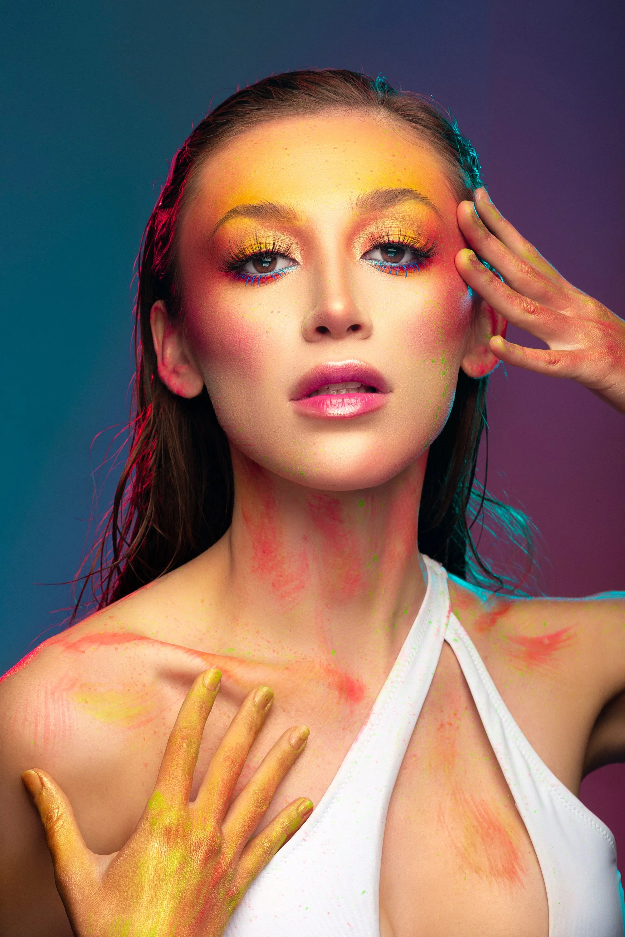 Retrato de una mujer con maquillaje colorido, cabello mojado, con pintura de colores en rostro, cuello y manos, con fondo de luz difusa de tonos fríos y cálidos.