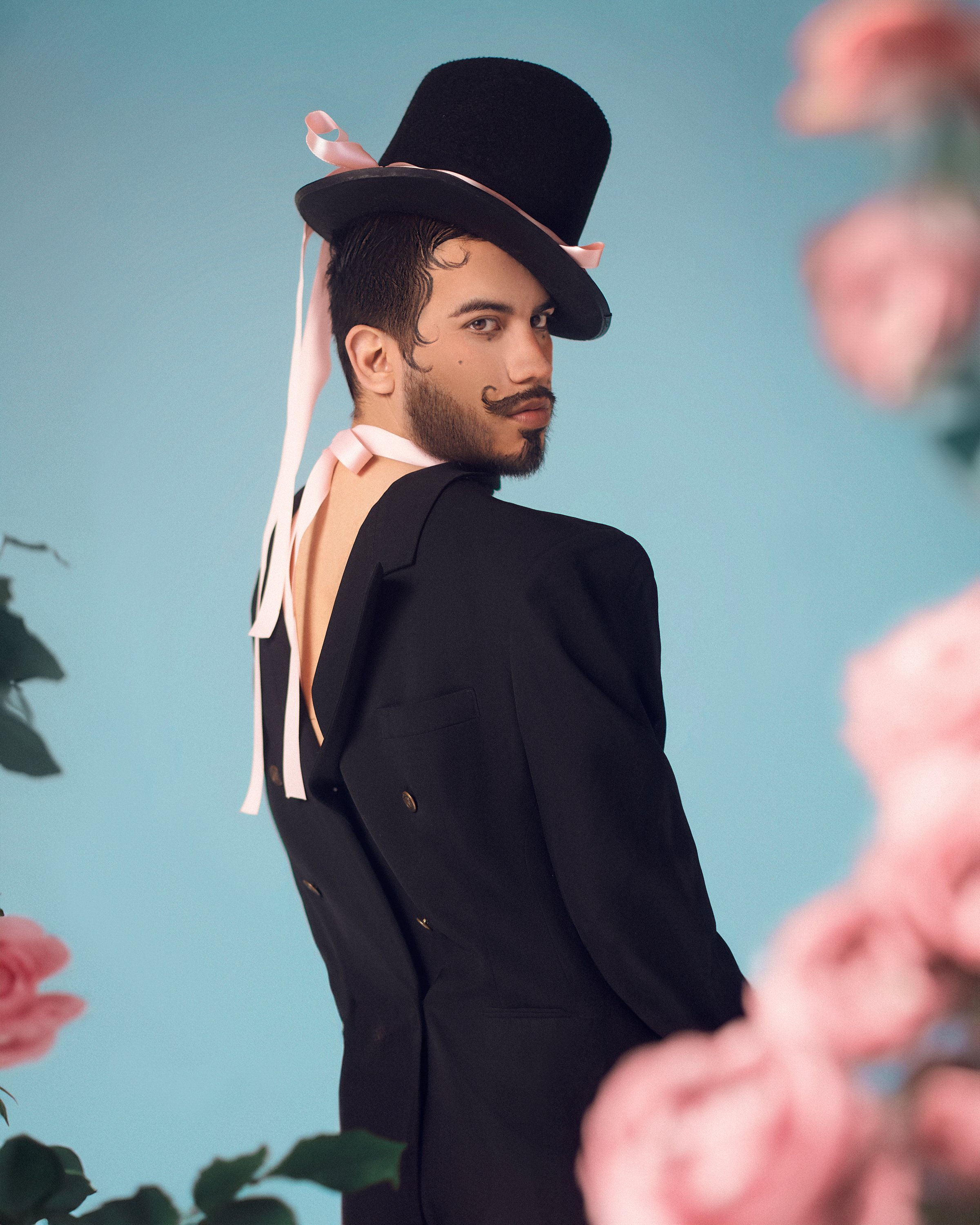 Hombre con barba y bigote, vestido con esmoquin negro, lleva un sombrero de copa negra con cinta rosa y lazo rosa, mirando de lado con fondo azul y flores rosas en el primer plano.