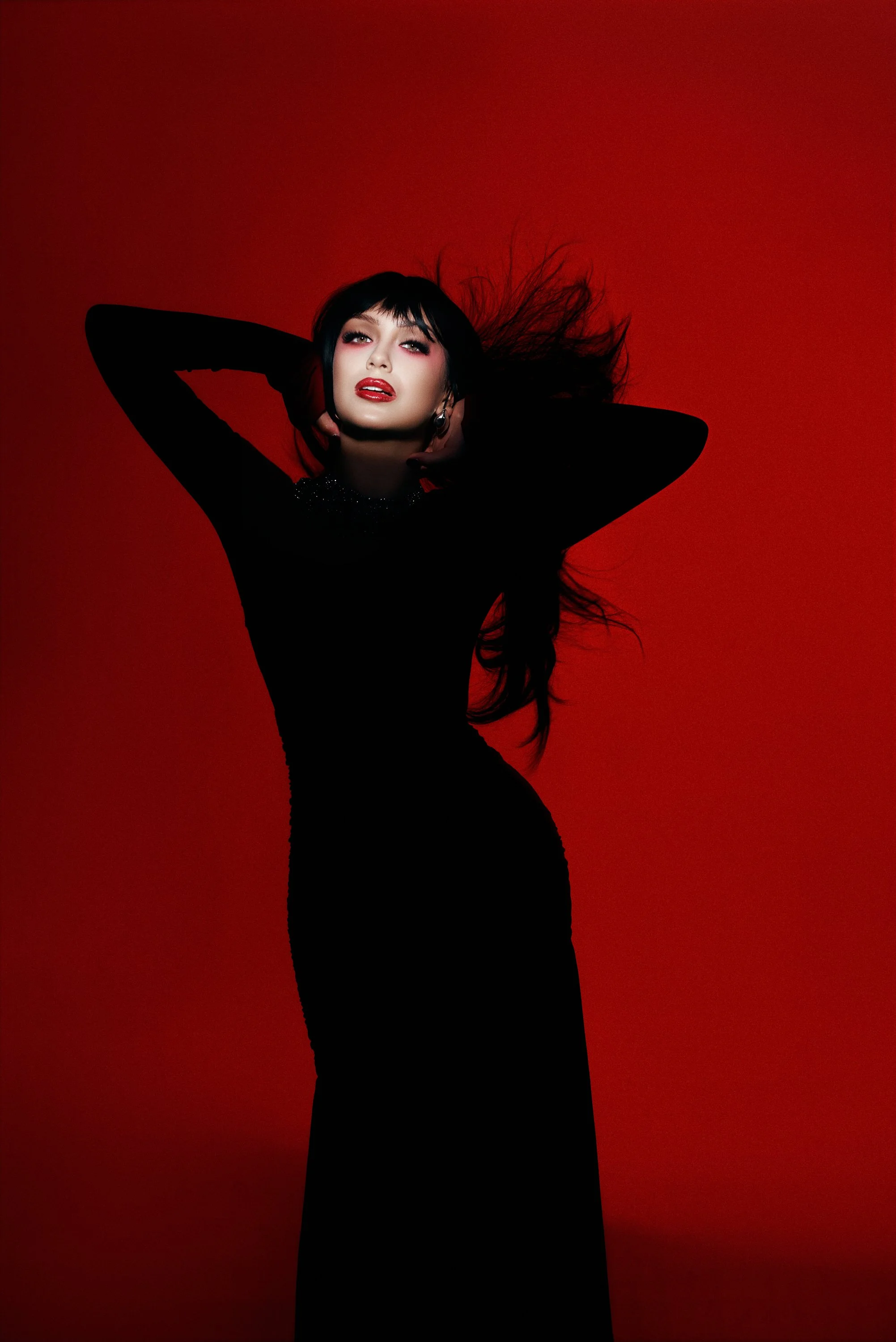 Mujer con cabello largo y oscuro, vistiendo un vestido negro ajustado y con fondo rojo, posando con las manos en la cabeza y expresión seductora.