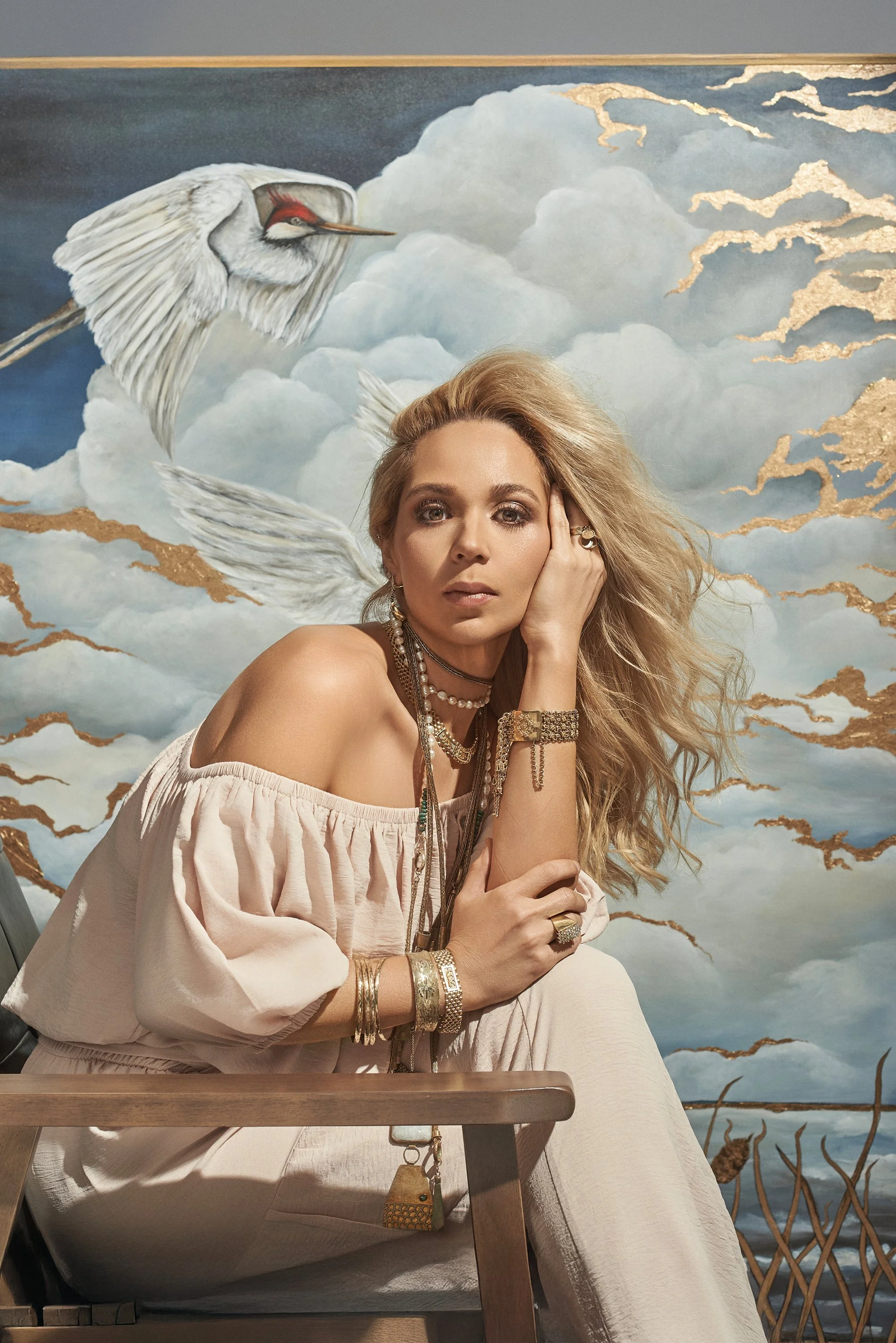 Mujer con cabello rubio y accesorios dorados, sentada con fondo de mural de nubes y un grulla.