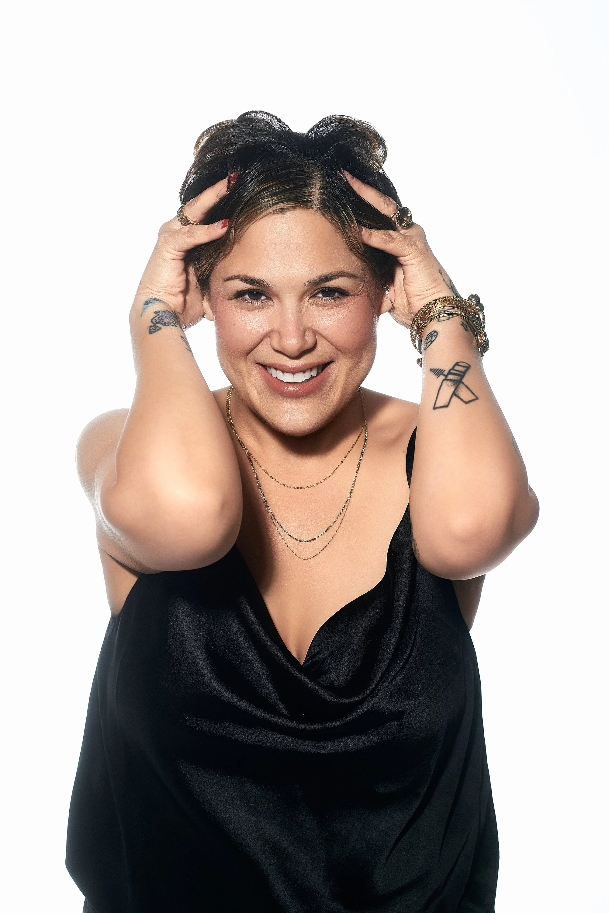 Mujer con cabello oscuro y tatuajes, usando vestido negro, sonriendo con las manos en el cabello, fondo blanco.