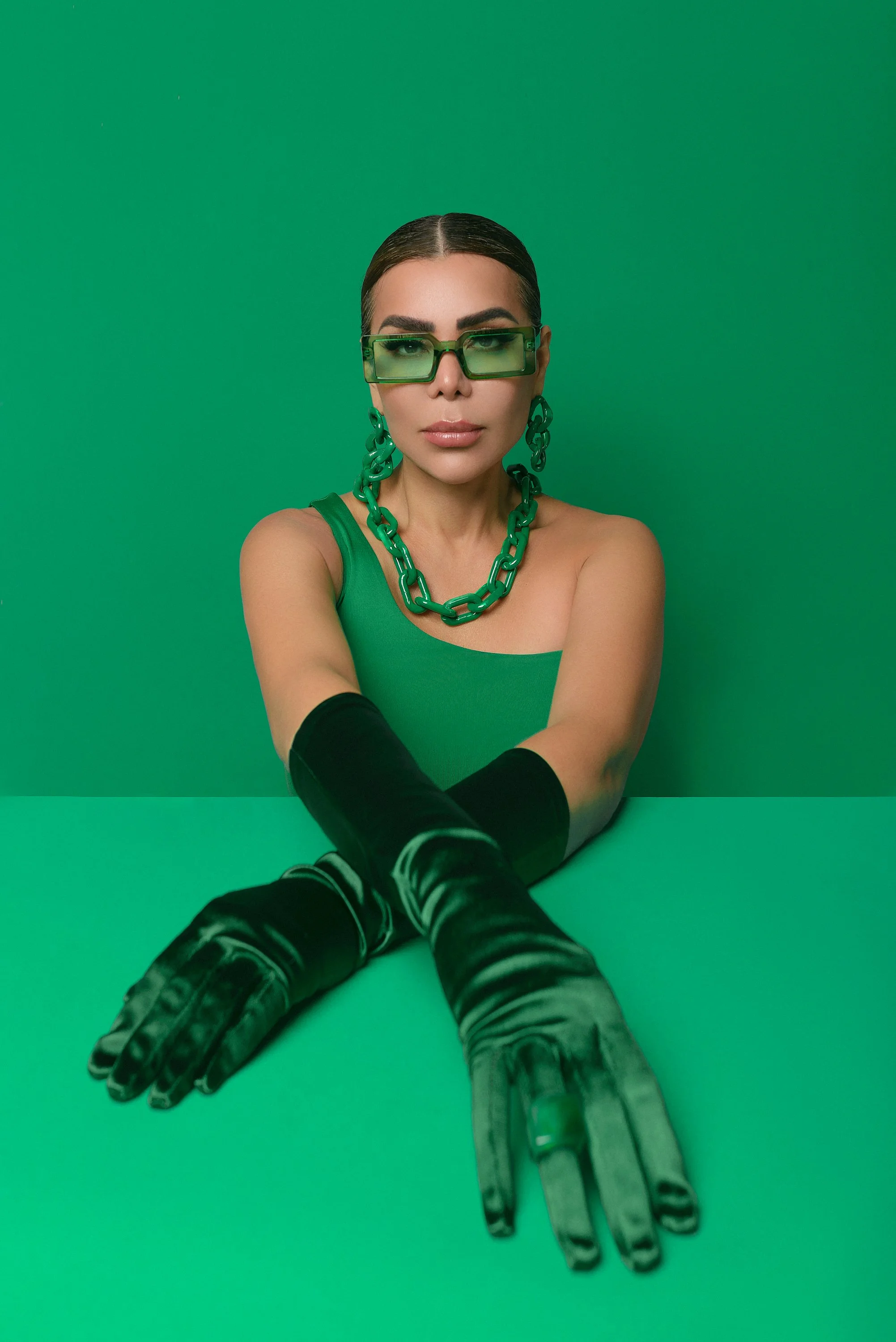 Mujer con gafas verdes, accesorios de cadena grande en color verde, vestido verde, guantes largos negros y fondo verde