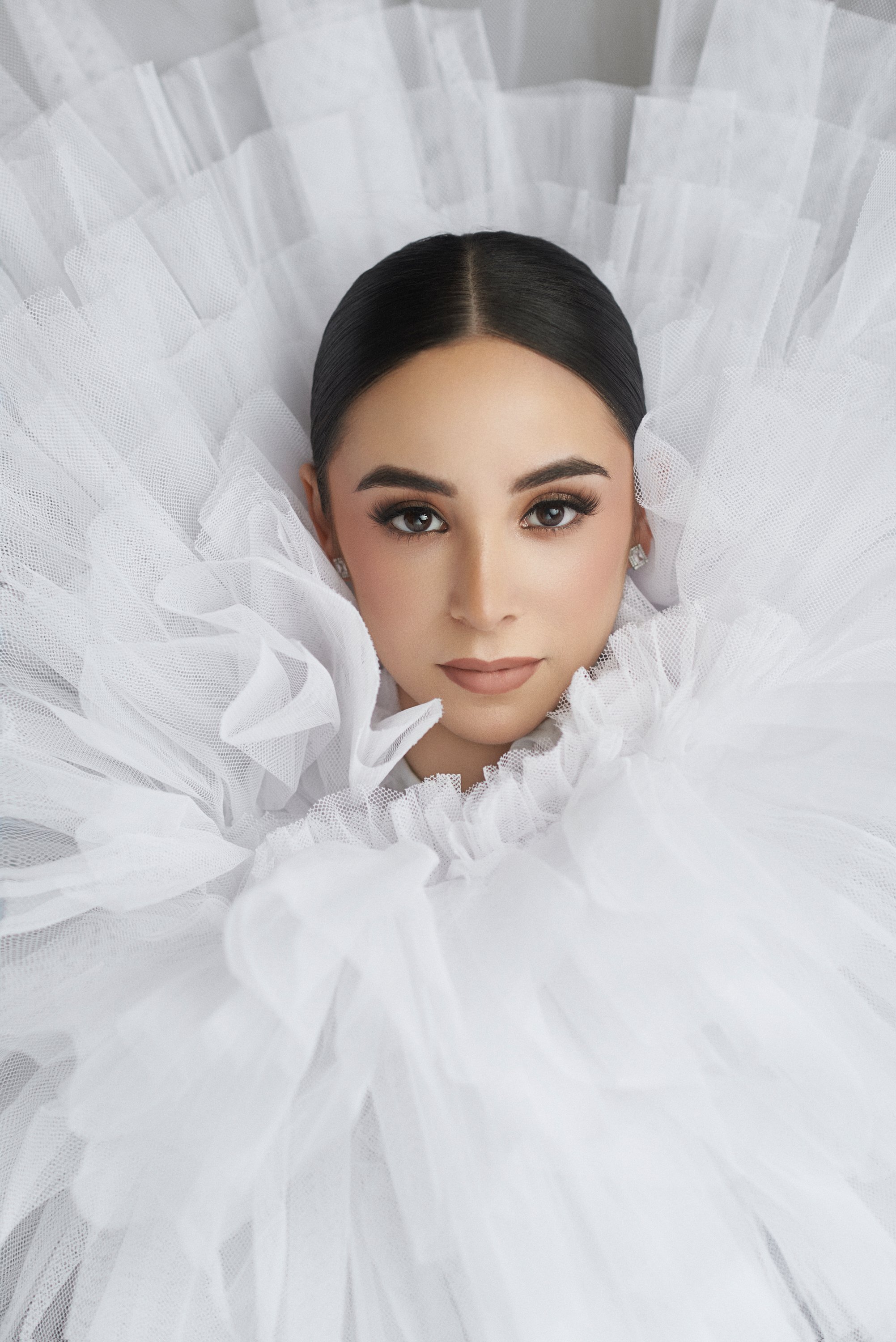 Retrato de una mujer con maquillaje, cabello liso y oscuro, rodeada de un vestido grande de tul blanco con volantes.