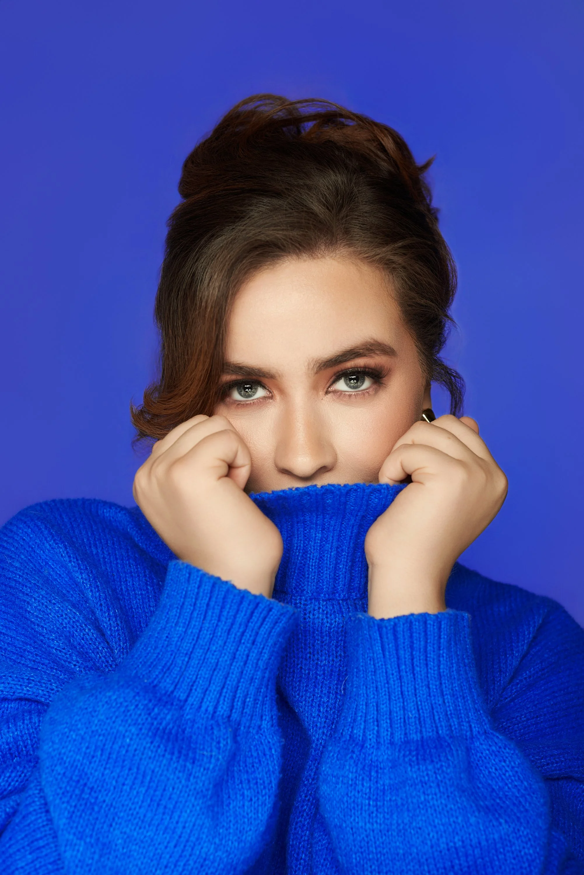 Mujer joven con ojos claros y cabello castaño, cubriéndose parcialmente la cara con un suéter azul, fondo azul sólido.