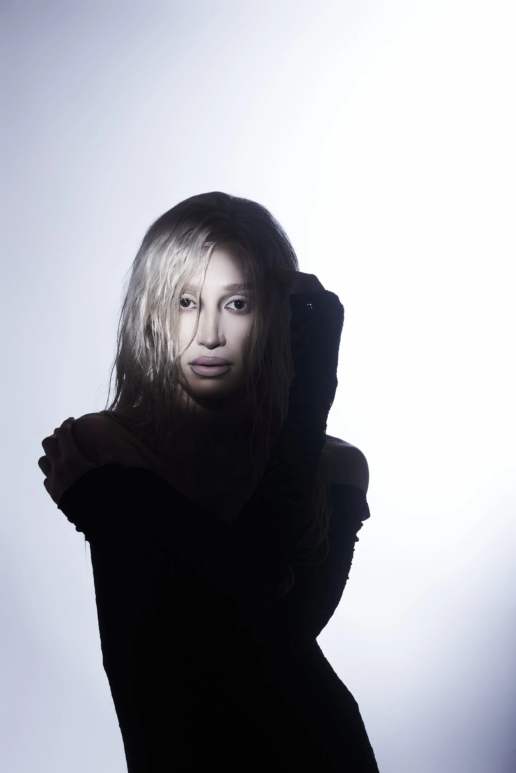 Mujer de cabello rubio y liso, con piel clara, vestido negro y guantes largos, posando frente a un fondo blanco con iluminación suave, mirando directamente a la cámara.