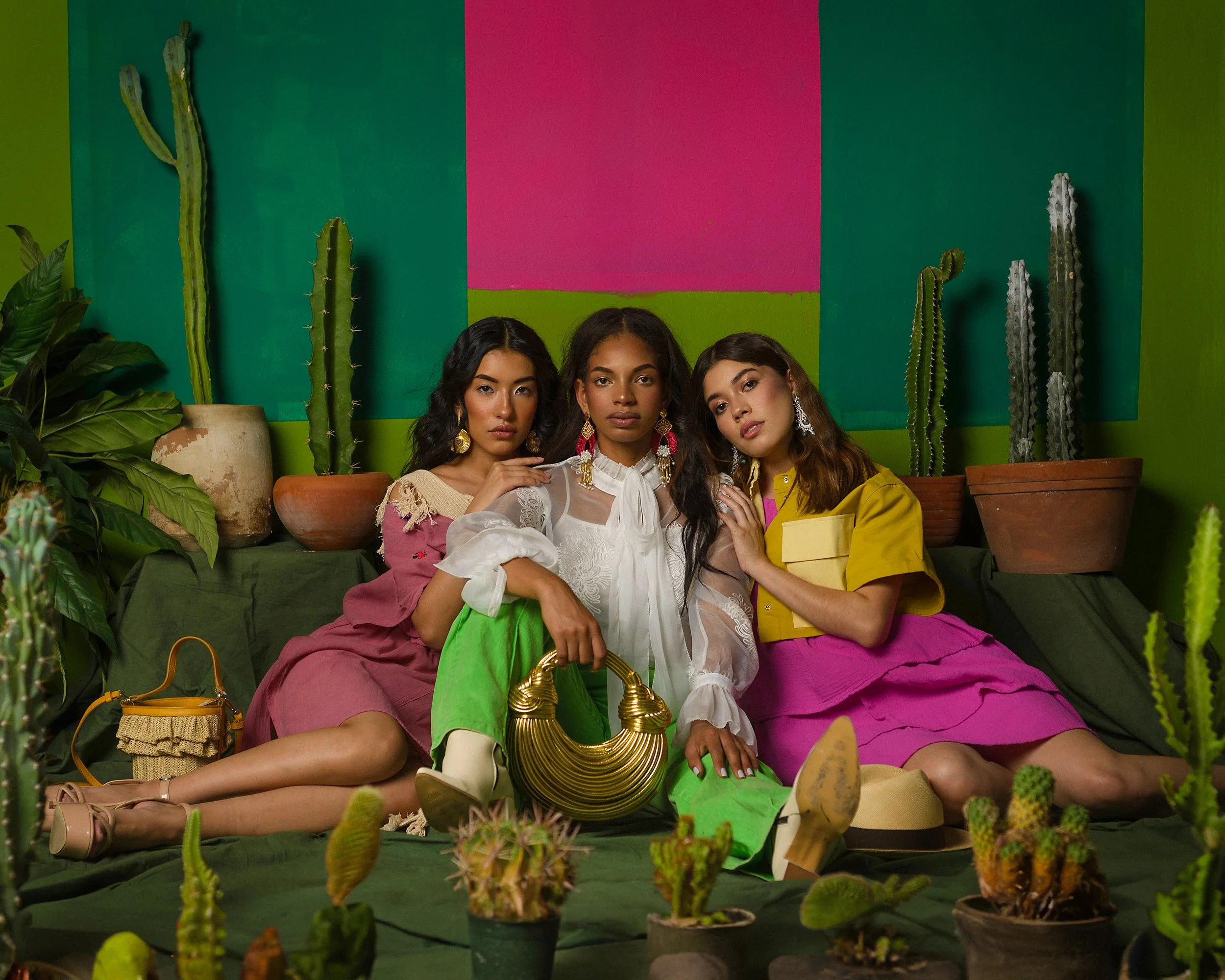 Tres mujeres sentadas en un ambiente decorativo con plantas de cactus en el fondo. Una lleva una blusa blanca con mangas de tul y pantalón verde, la otra usa un top rosa y falda rosa, y la otra una blusa amarilla con falda rosa. Están rodeadas de pla