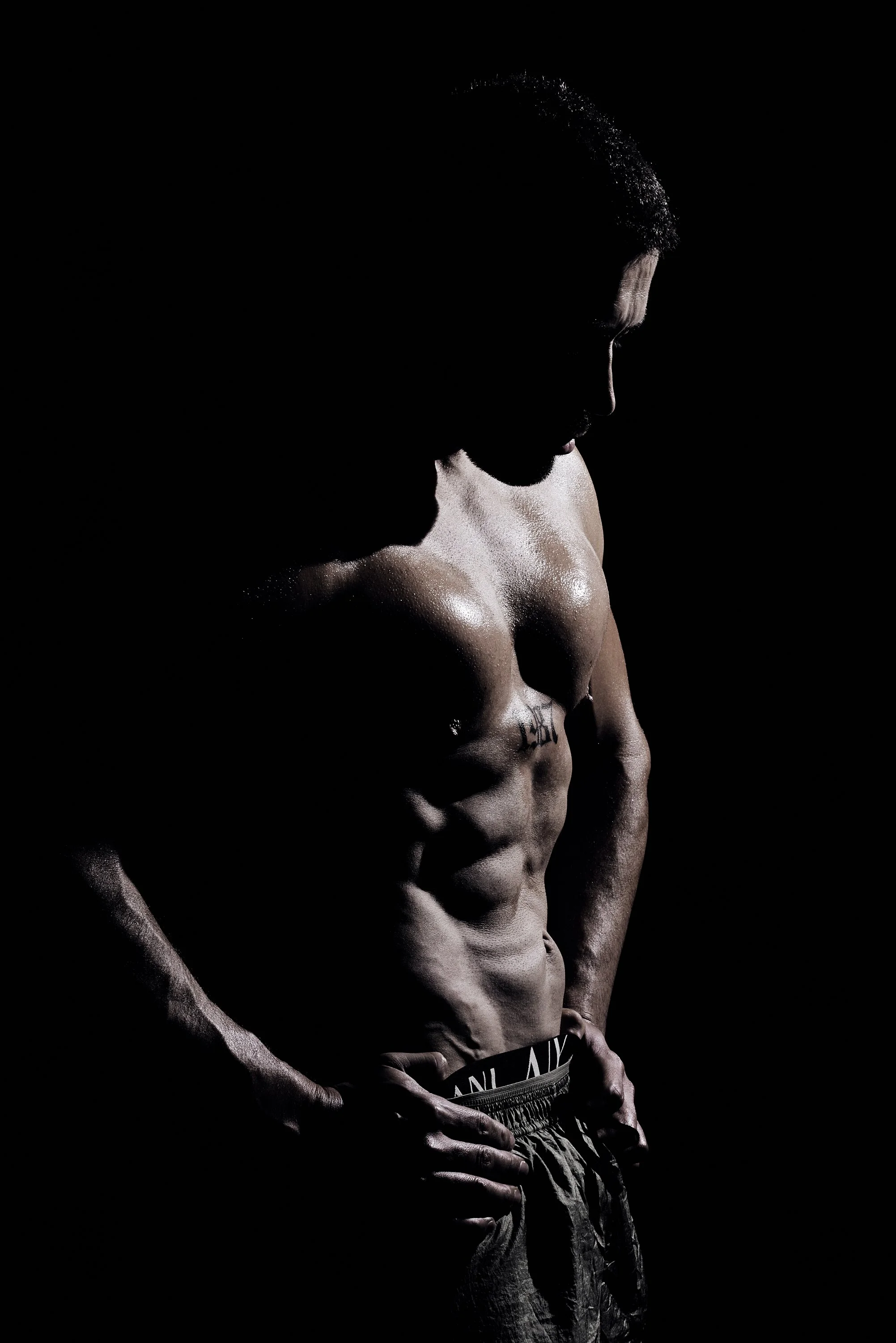 Hombre musculoso sin camisa, de perfil, con las manos en las caderas, en un fondo negro, con luz que resalta sus músculos abdominales y pectorales.