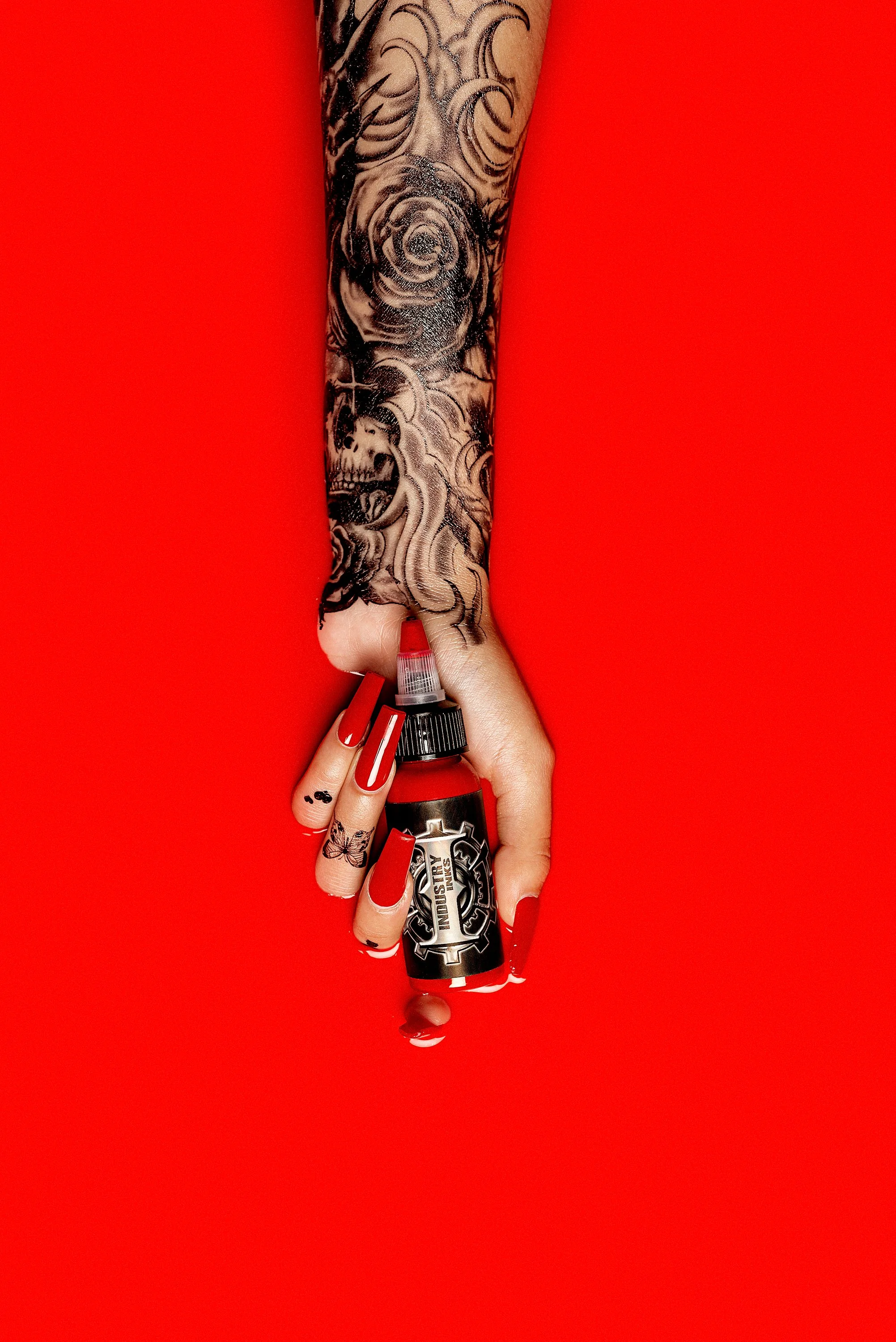 Brazo con tatuajes sosteniendo un frasco de tinta de dar color a tatuajes con fondo rojo.