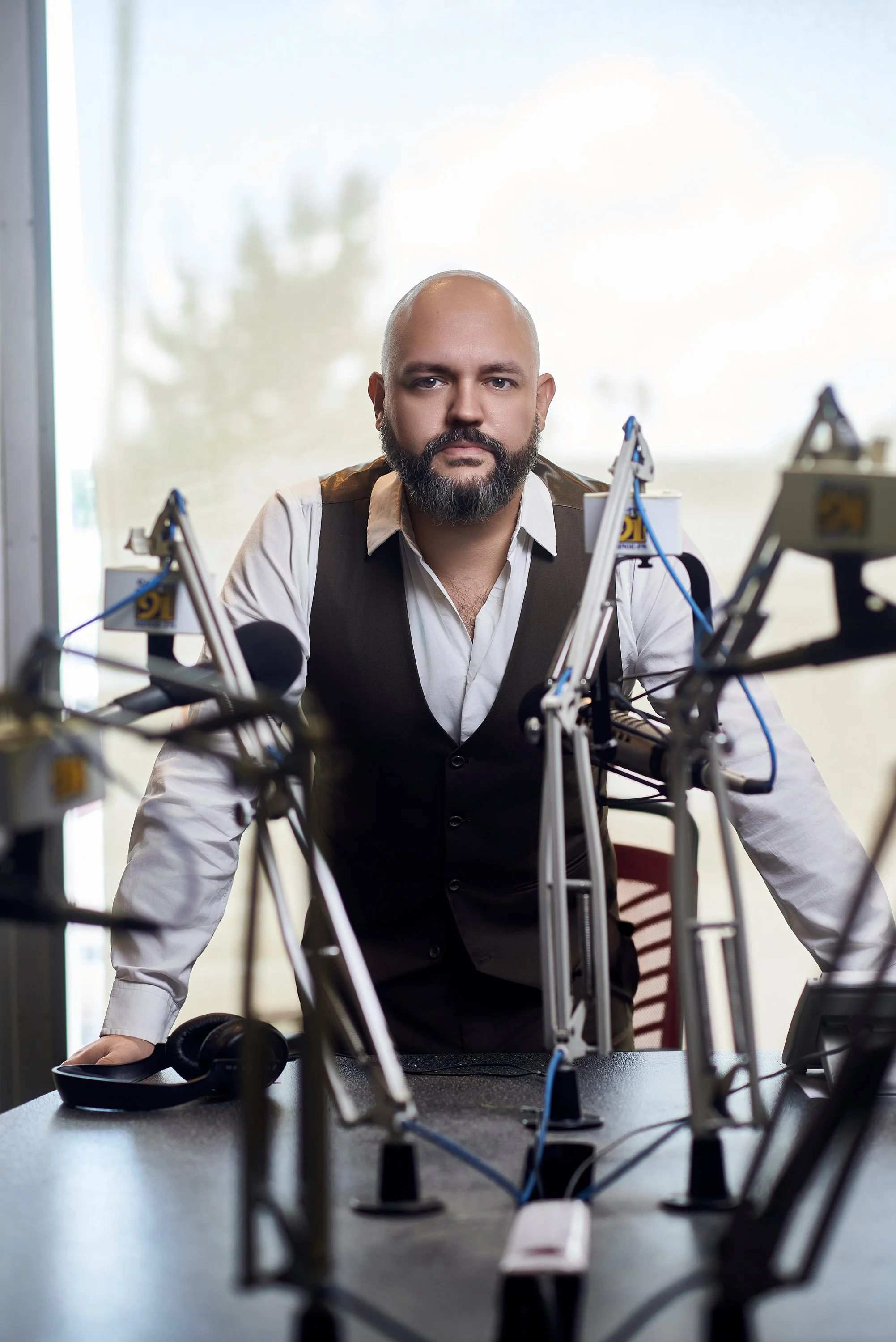 Un hombre con barba y cabello calvo, vestido con camisa blanca y chaleco negro, se encuentra frente a varias prótesis o dispositivos tecnológicos en un laboratorio o taller.