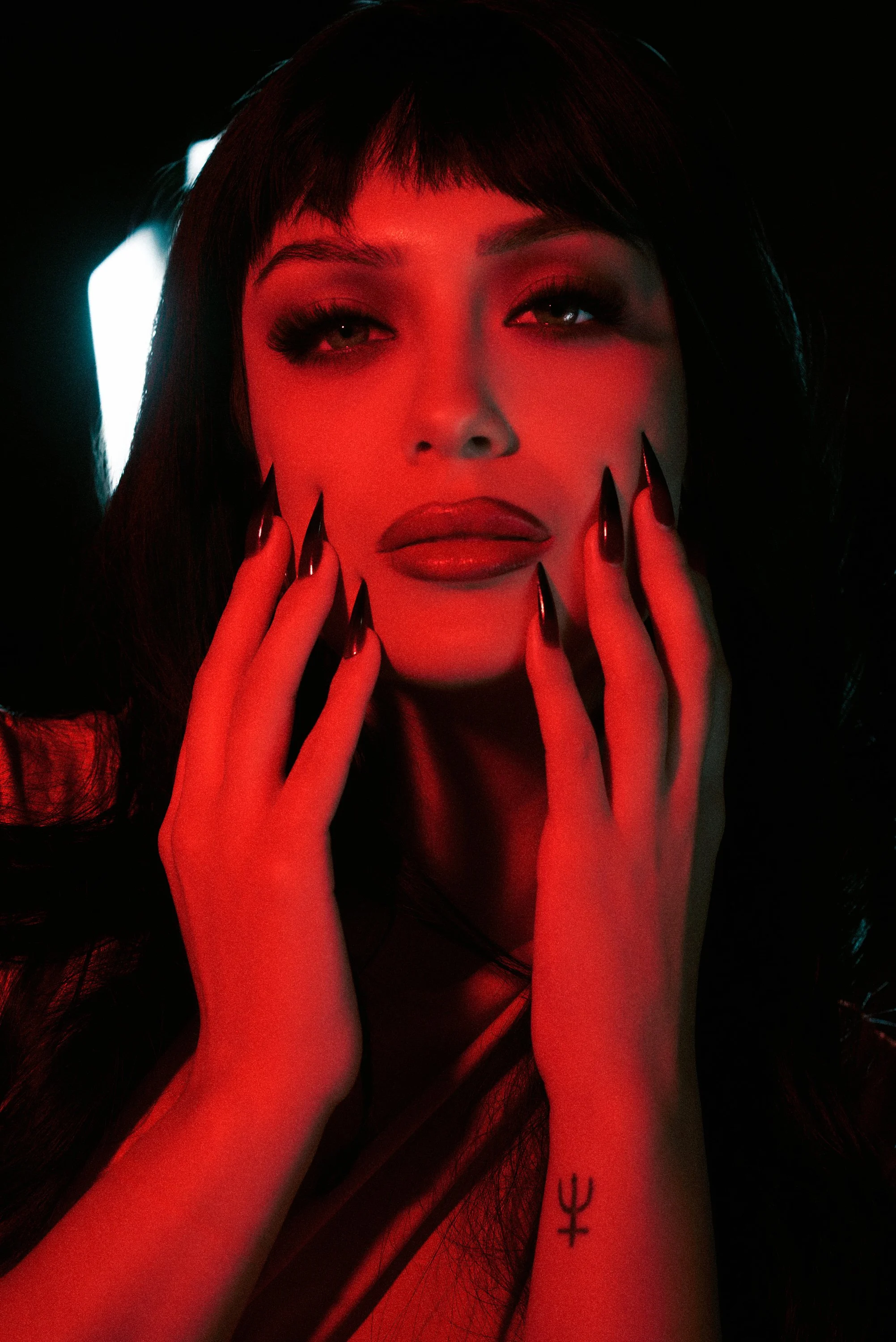 Retrato de una mujer con maquillaje llamativo, uñas largas y negras, y un símbolo en la muñeca, en un entorno con iluminación de luz roja y fondo oscuro.