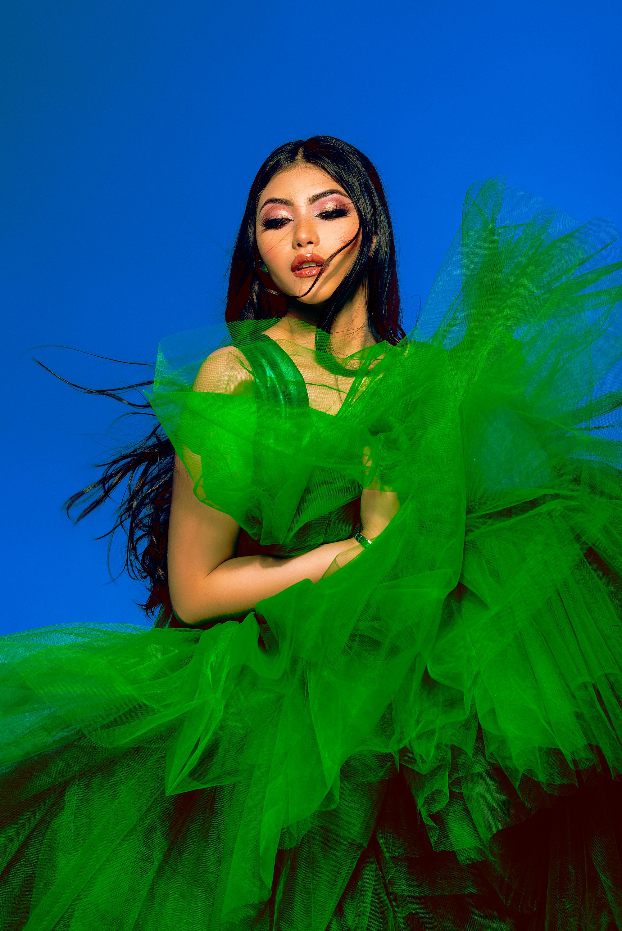 Mujer con vestido de tul verde y maquillaje brillante posando contra un cielo azul.