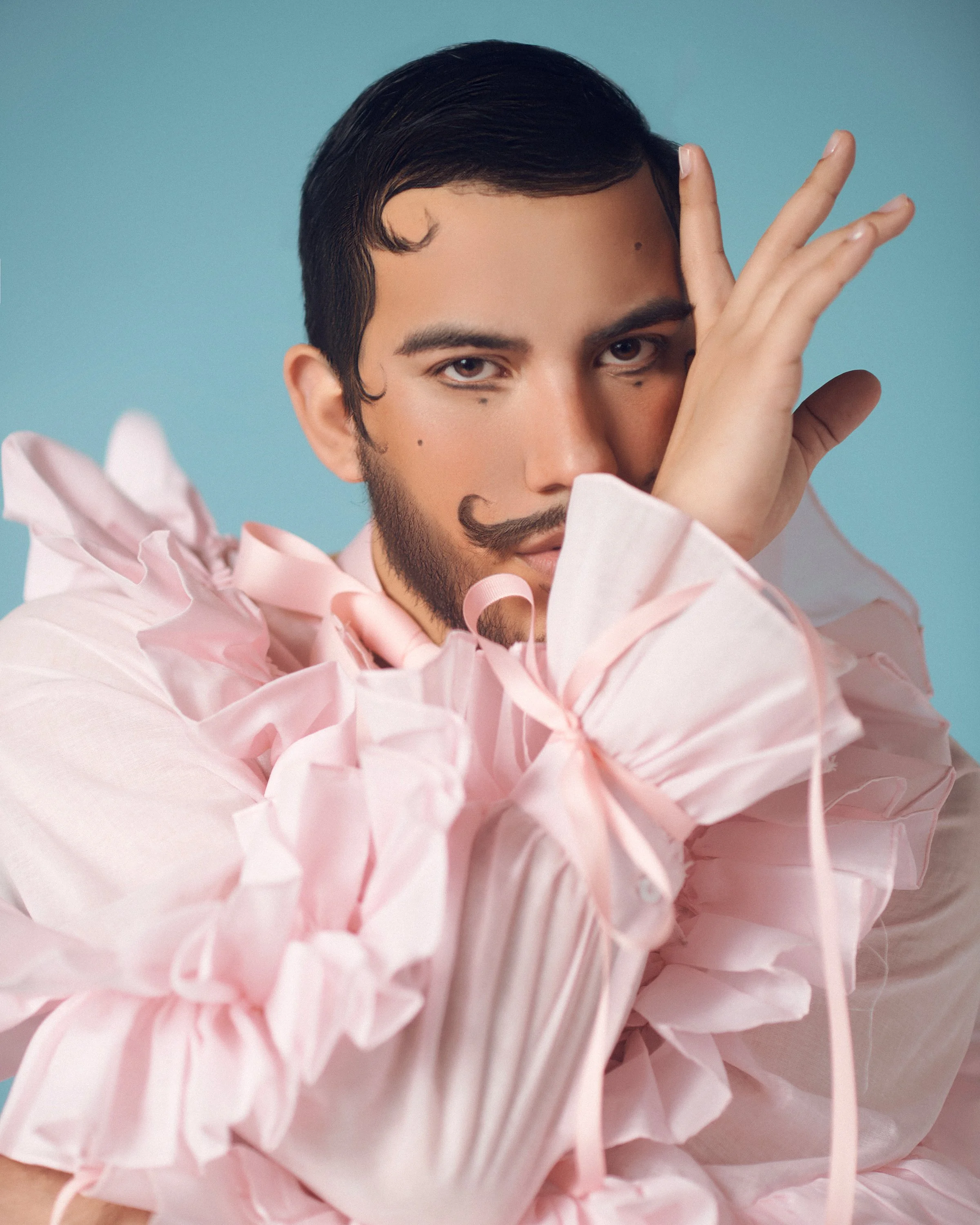 Joven con barba, bigote y cabello oscuro peinado hacia un lado, con una expresión artística, lleva ropa con muchas volantes rosados y frente a un fondo azul claro.