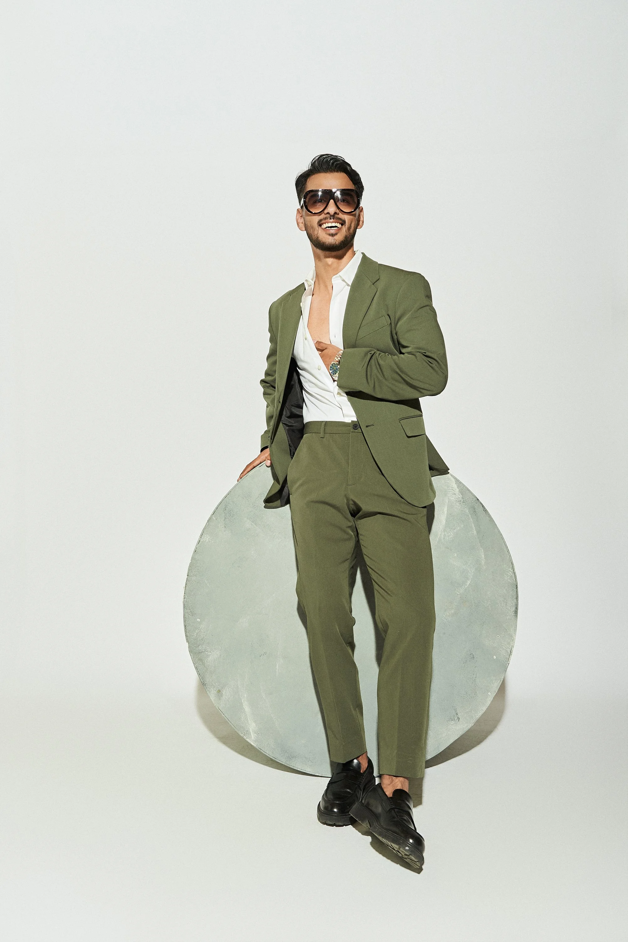 Hombre con traje verde, sandalias negras, gafas de sol oscuras, sonriendo, posando con fondo blanco y una luna de cartón grandes detrás de él.