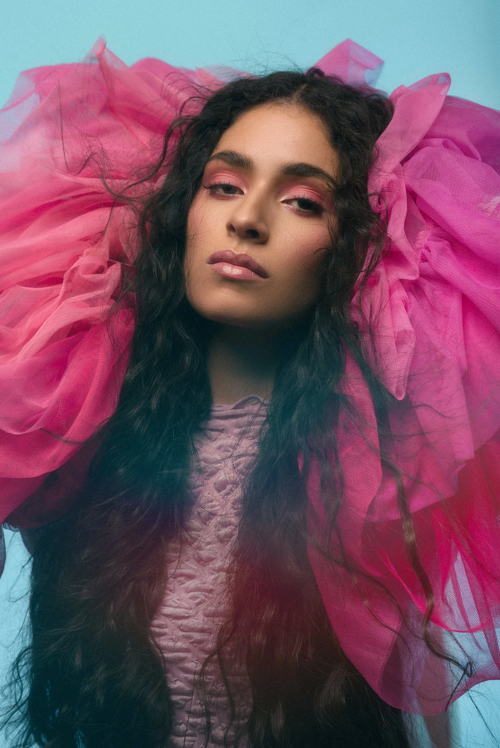 Mujer con piel clara y cabello rizado oscuro, usando maquillaje rosa y ropa de tela rosa con volumen, sobre fondo azul.