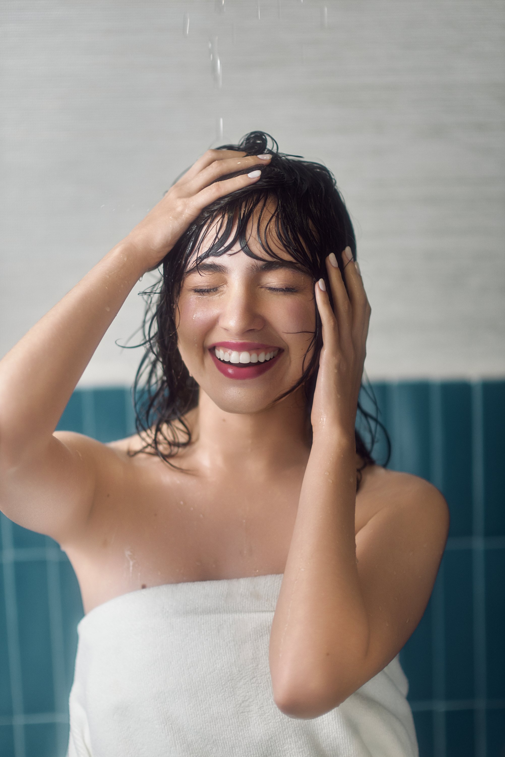 Mujer sonriendo y disfrutando en la ducha, con cabello mojado y tocándose la cabeza y la cara.