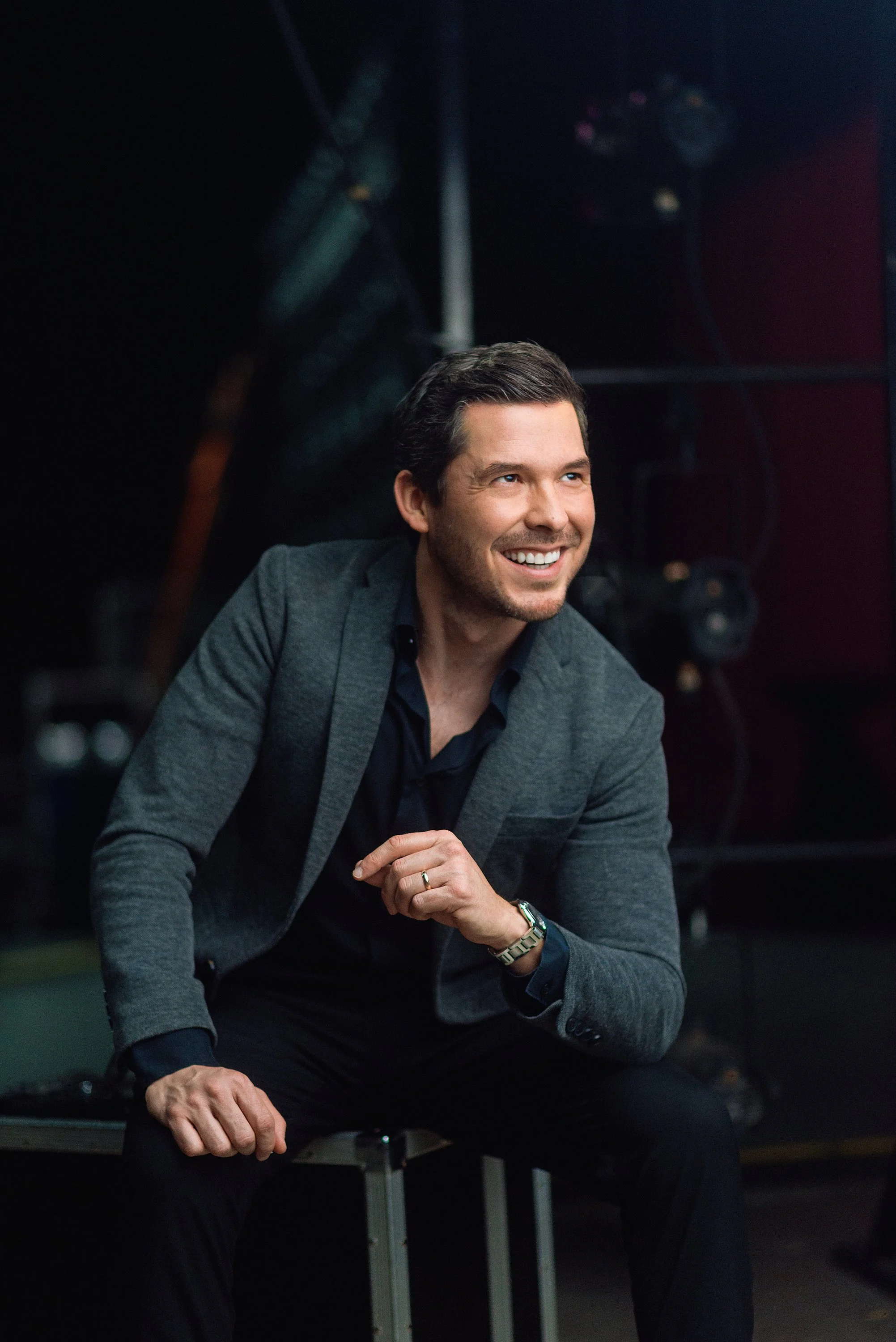 Hombre sonriendo y sentado en un banco, vestido con una chaqueta gris y camisa negra, en un entorno oscuro con equipo de teatro o escenario al fondo.