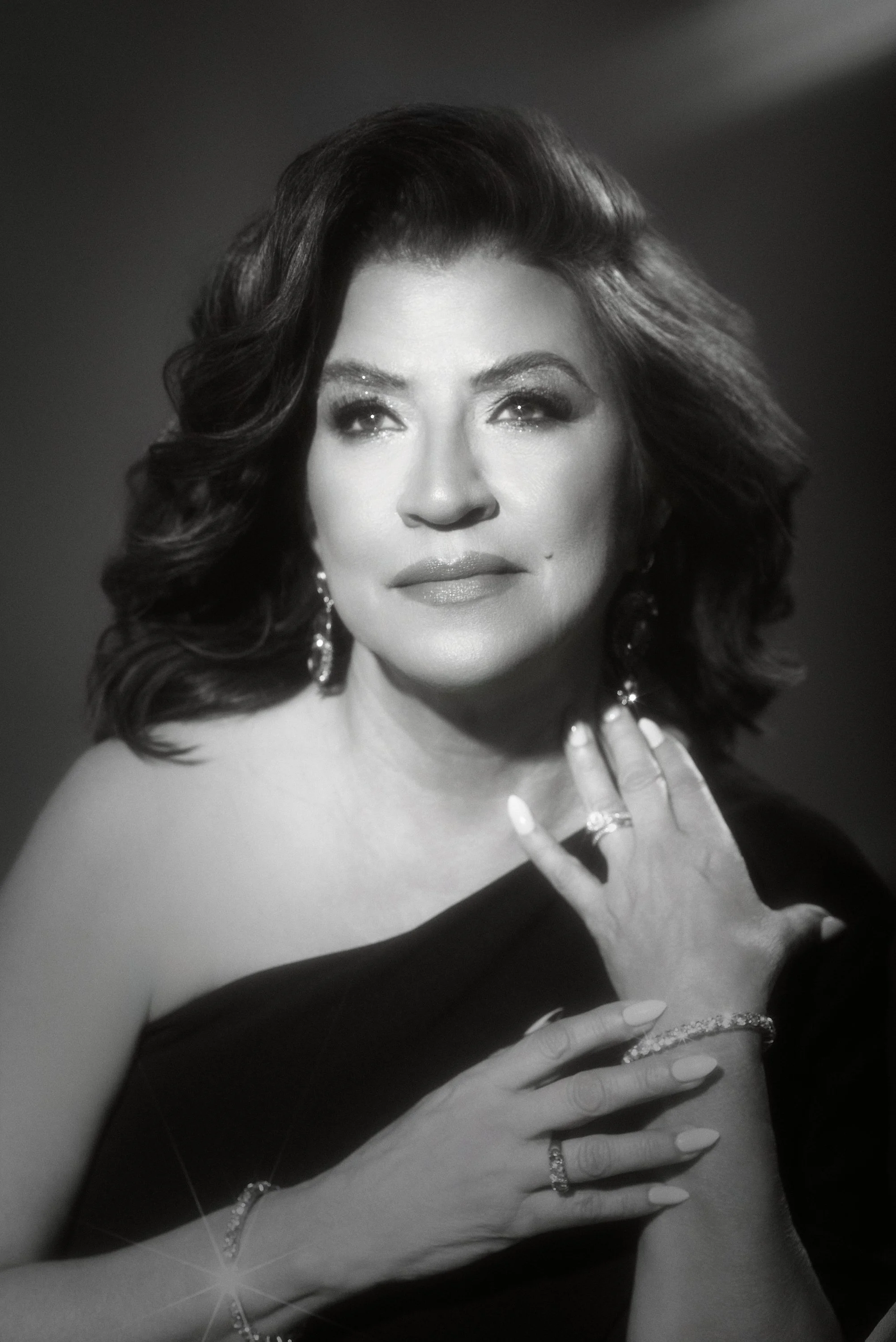 Retrato en blanco y negro de una mujer con cabello rizado y peinado vintage, usando joyería elegante, con una expresión de serenidad y mirada en dirección ligeramente hacia arriba, con las manos levantadas y adornadas con pulseras y anillos.