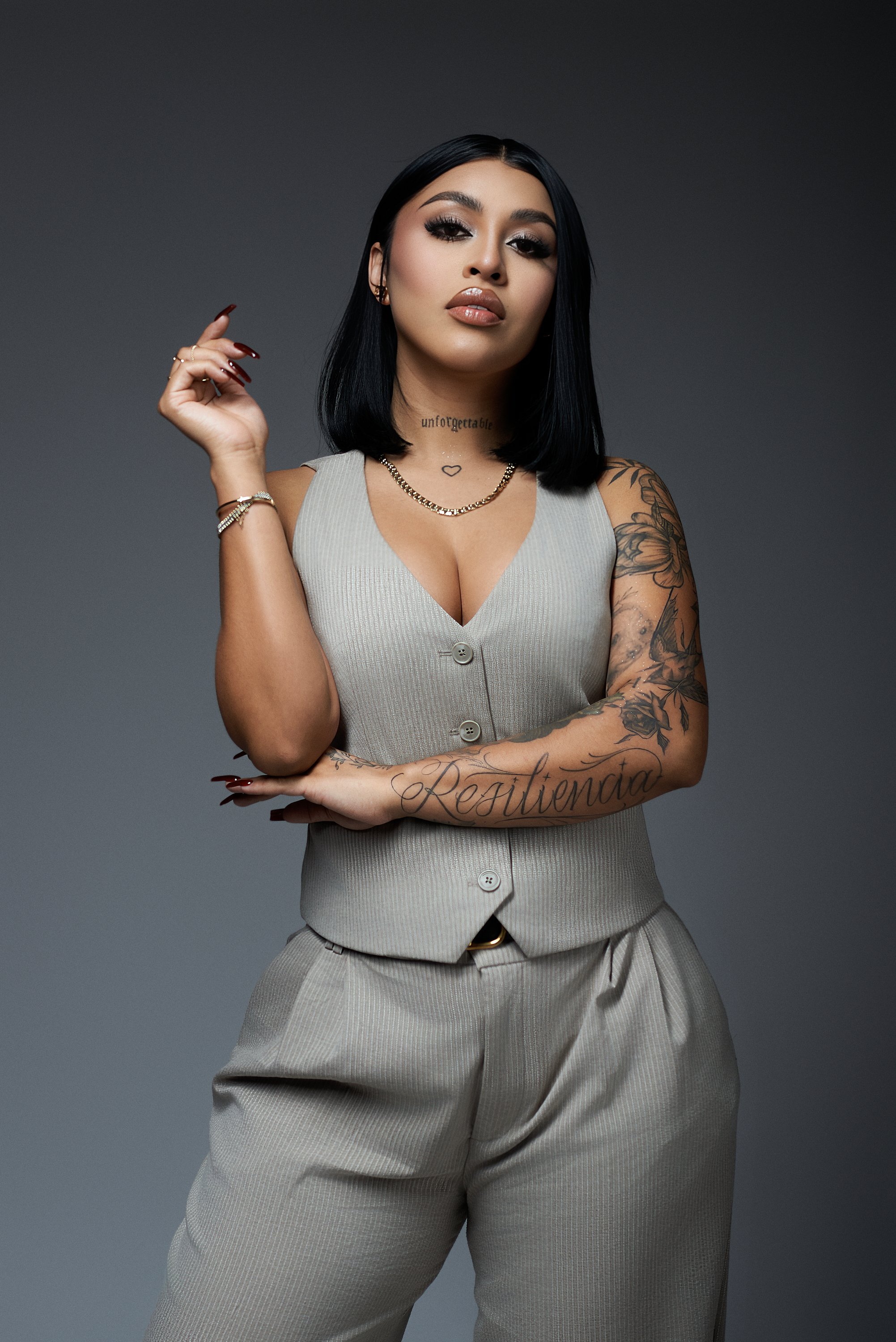 Retrato de una mujer con cabello negro, vestida con un conjunto beige, con tatuajes en los brazos y el cuello, posando con expresión segura contra un fondo gris oscuro.