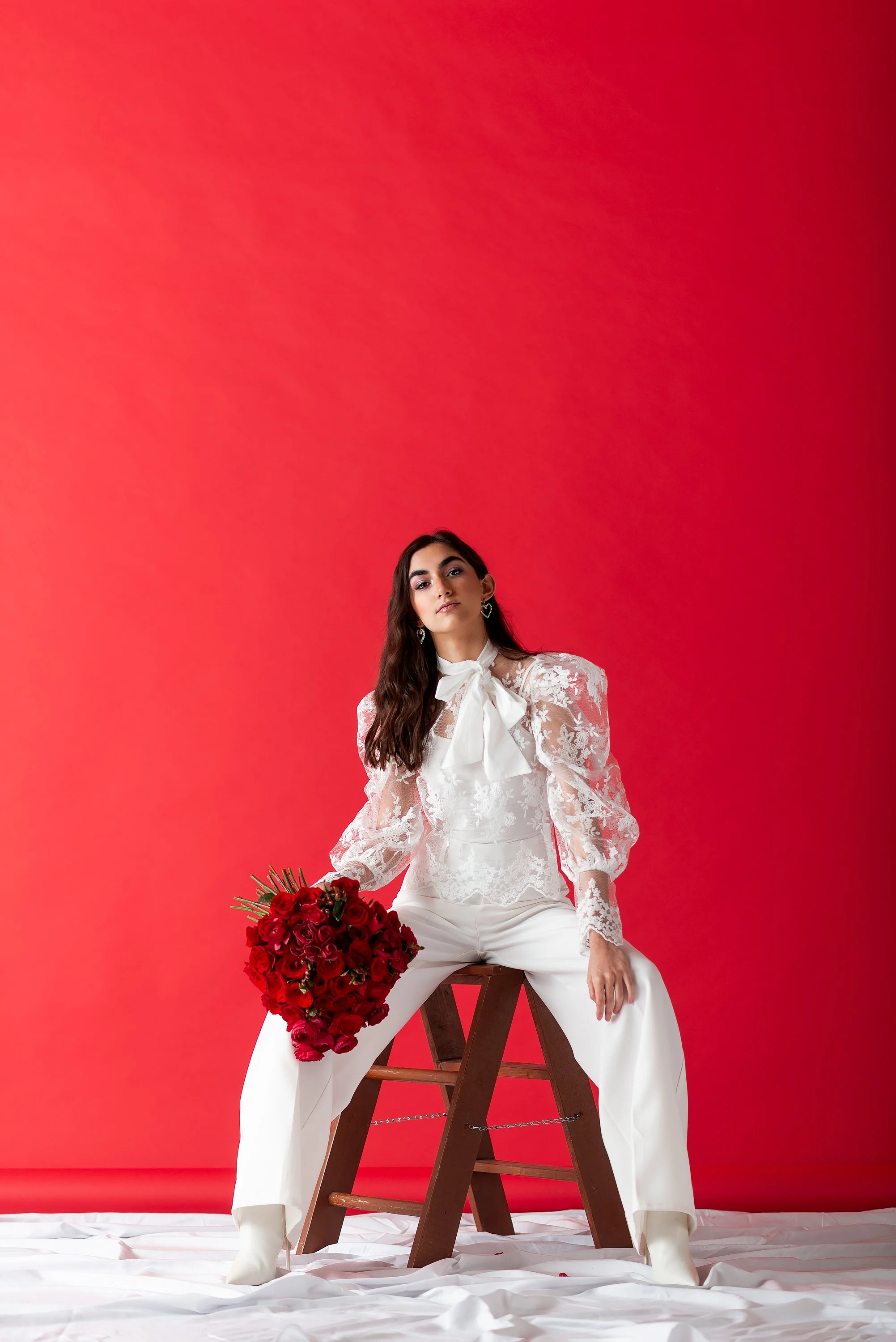 Mujer vestida con blusa blanca de encaje y pantalones blancos, sentada en un taburete de madera, sosteniendo un ramo de rosas rojas, con fondo rojo y suelo cubierto con tela blanca.