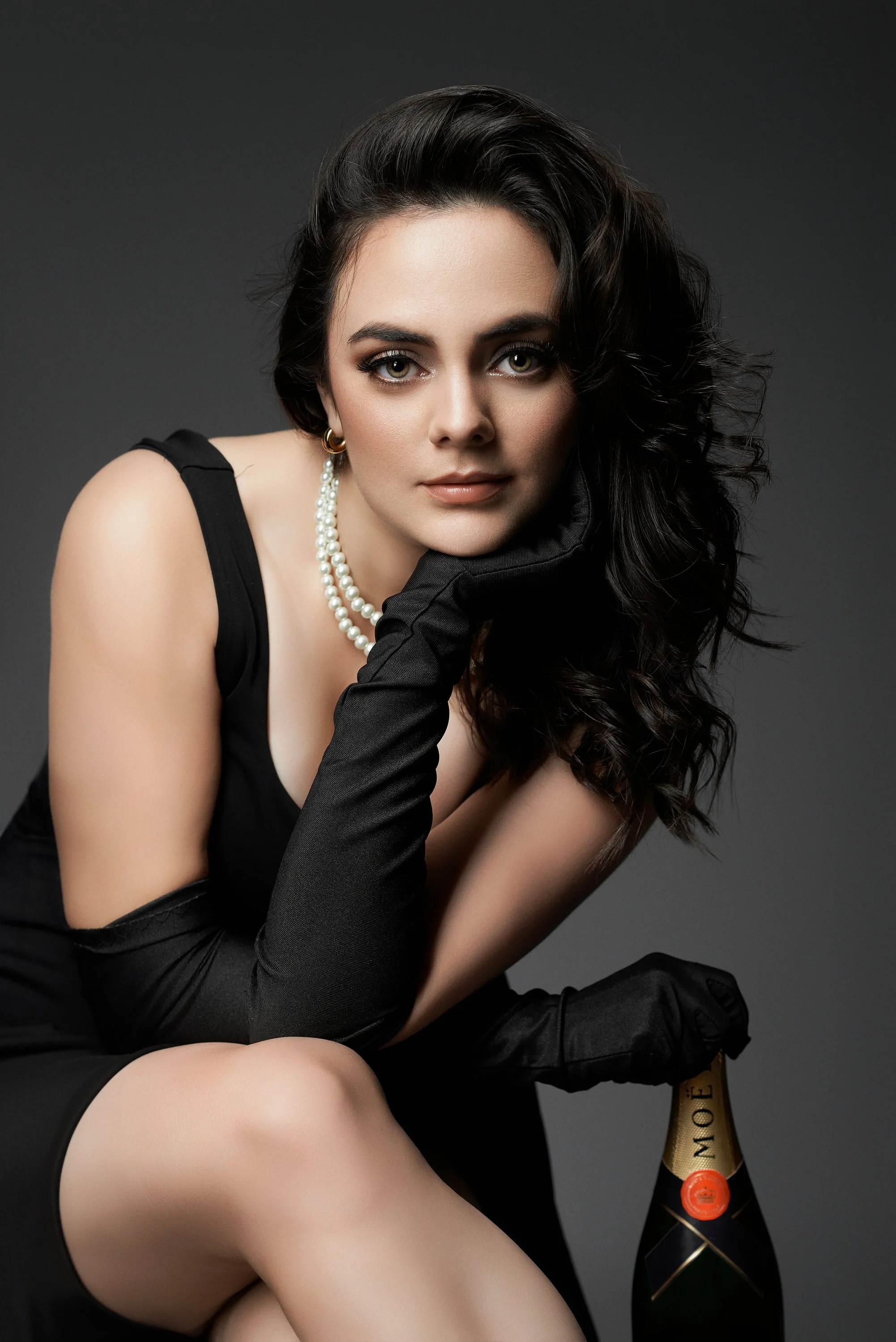 Mujer con cabello rizado, vestido negro, guantes negros largos, perlas, sosteniendo una botella de champagne Moët y con expresión pensativa.