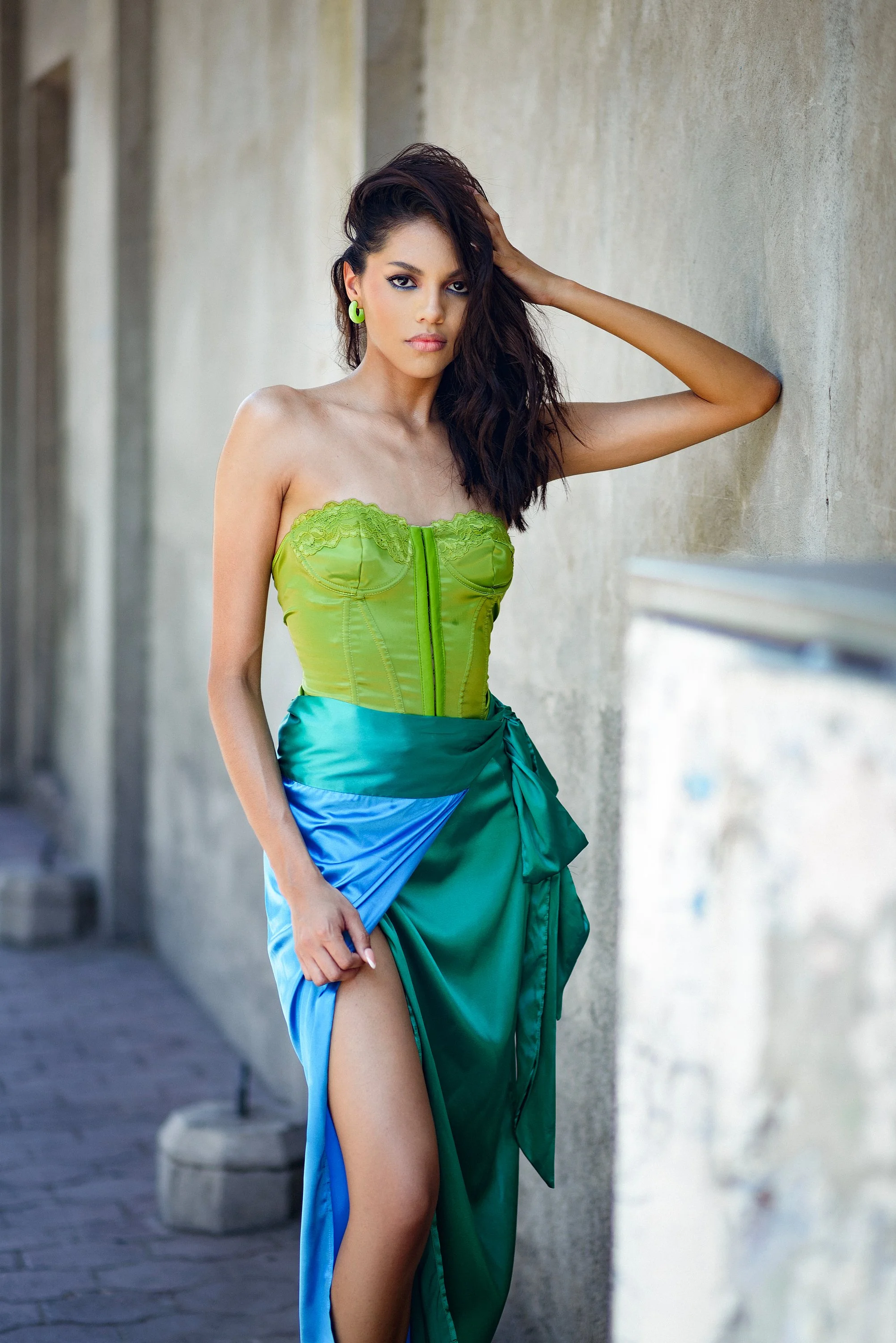Mujer con vestido corto en el centro, con falda larga y abertura, posando junto a una pared de concreto, con cabello rizado y oscuro, en un entorno urbano.