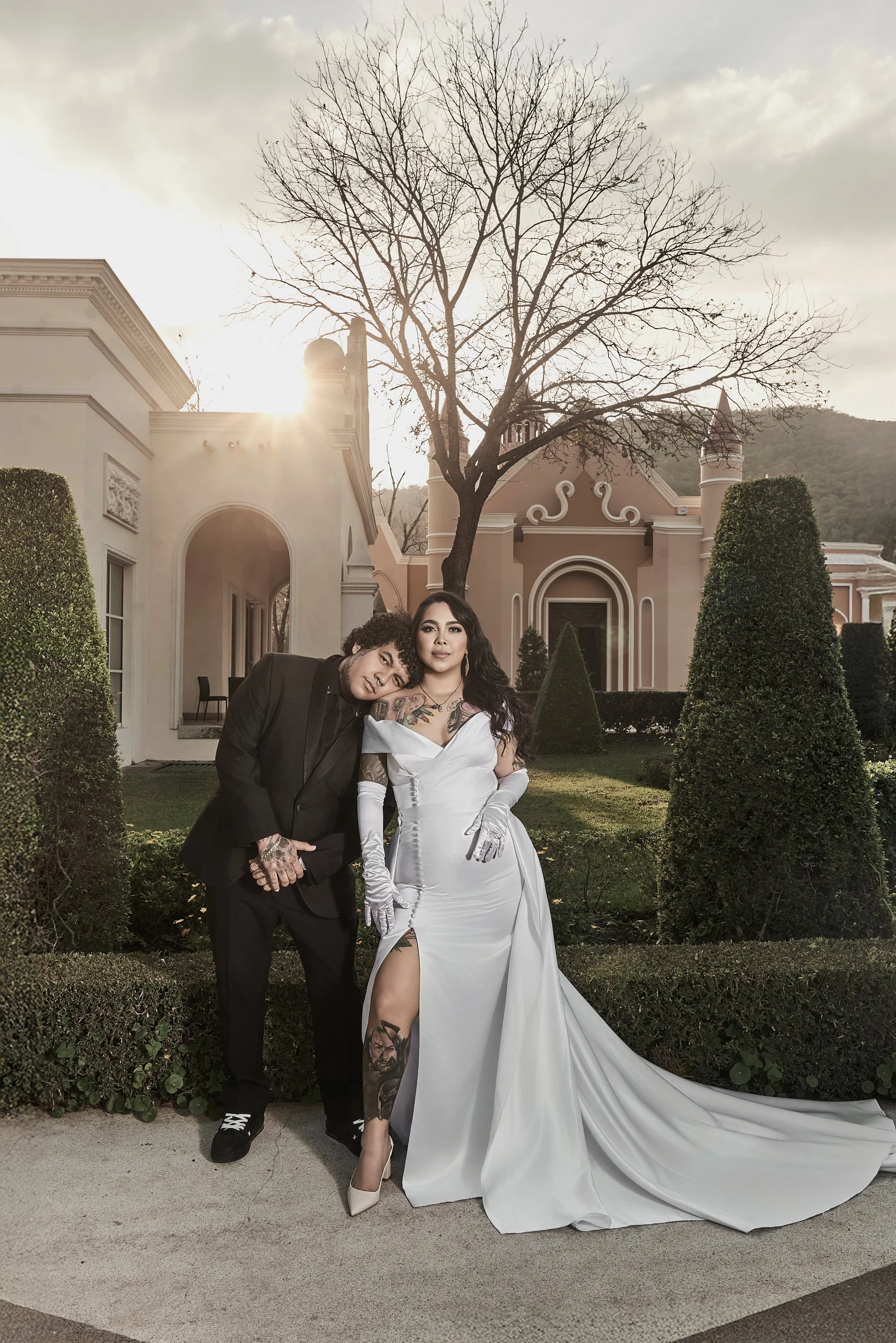 Pareja en atuendo formal, mujer en vestido blanco largo y hombre en traje negro, posando en un jardín con árboles y una casa elegante al fondo, al atardecer.