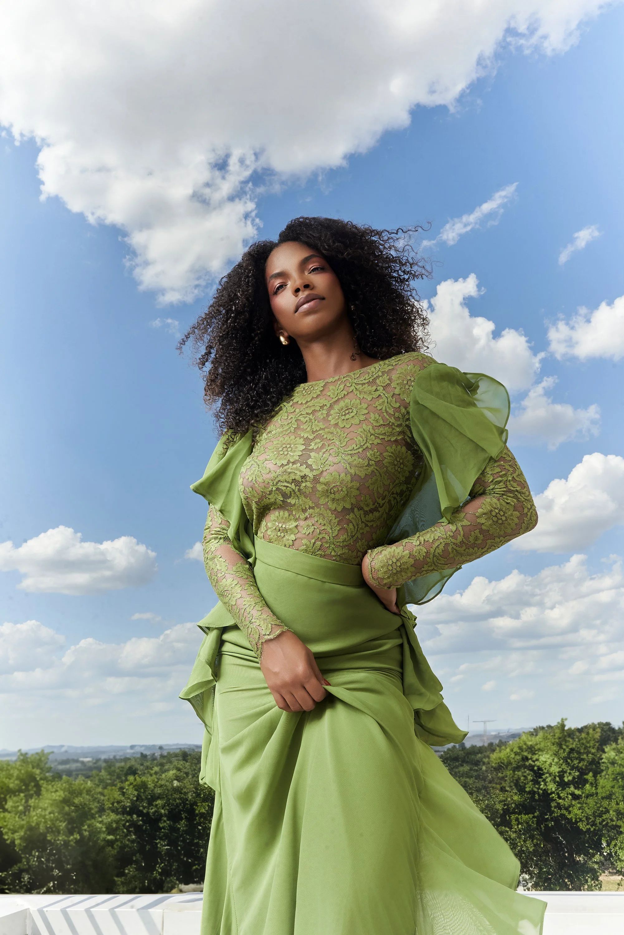 Mujer con vestido verde con encaje y detalles en tul, posando en un espacio abierto con cielo y nubes.