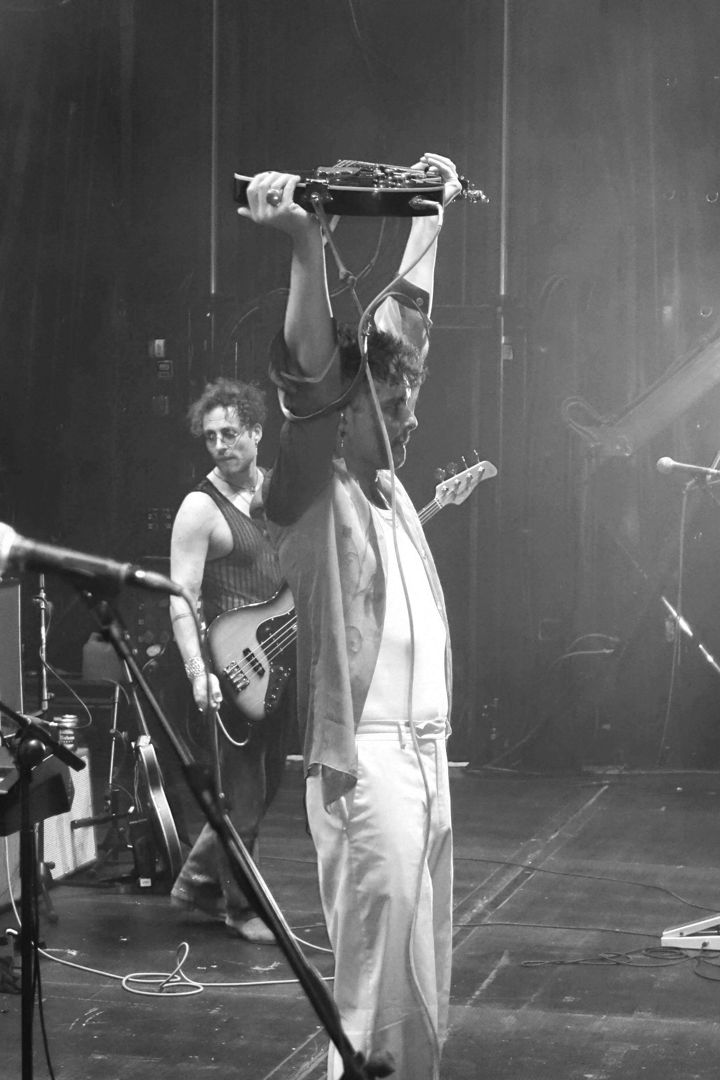 A black and white photo of a musician holding a guitar above their head on stage, with another band member playing a bass guitar in the background. Stage equipment and cables are visible.